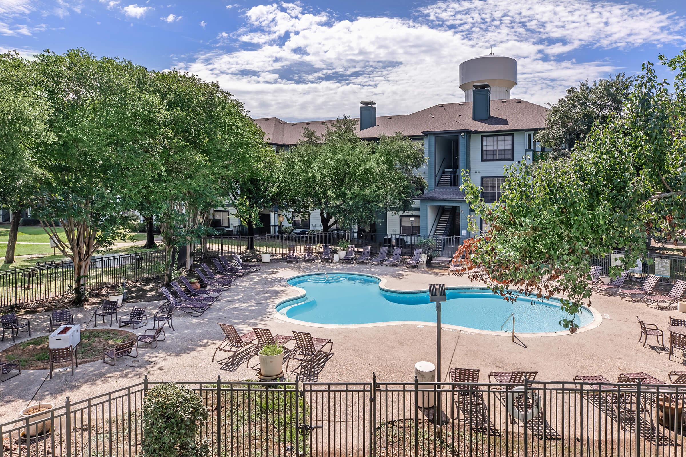 A serene outdoor scene featuring a swimming pool surrounded by lounge chairs and greenery. In the background, there are apartment buildings under a partly cloudy sky. The setting is well-maintained and inviting, ideal for relaxation and leisure.