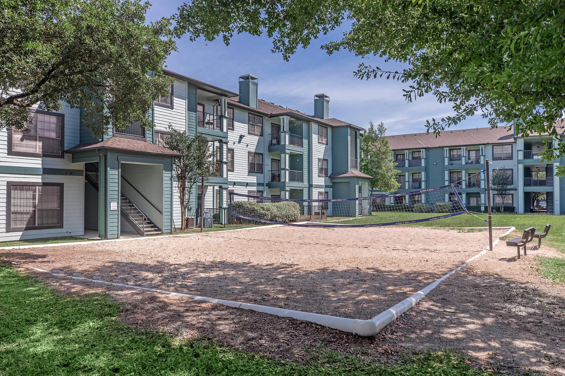 A sandy volleyball court with a net, surrounded by green grass and trees, located in front of two apartment buildings. The buildings have a modern design with multiple windows and balconies, and the sky is clear blue with a few clouds. A bench is positioned nearby for relaxation.