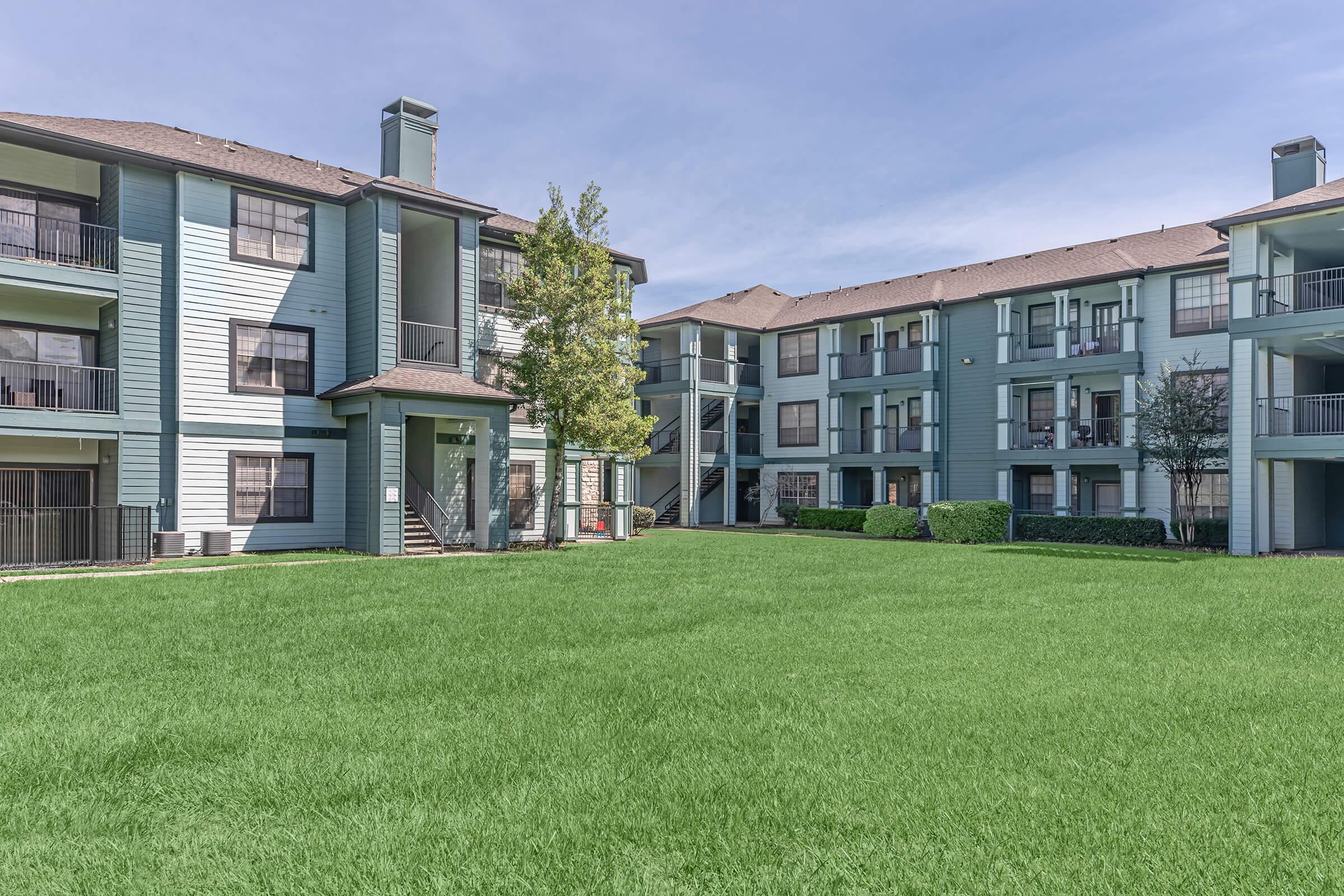 View of a residential apartment complex featuring two multi-story buildings with blue-green exteriors. A well-maintained lawn with lush green grass is in the foreground, and trees are seen between the buildings, providing a pleasant outdoor area for residents. The sky is clear and bright.