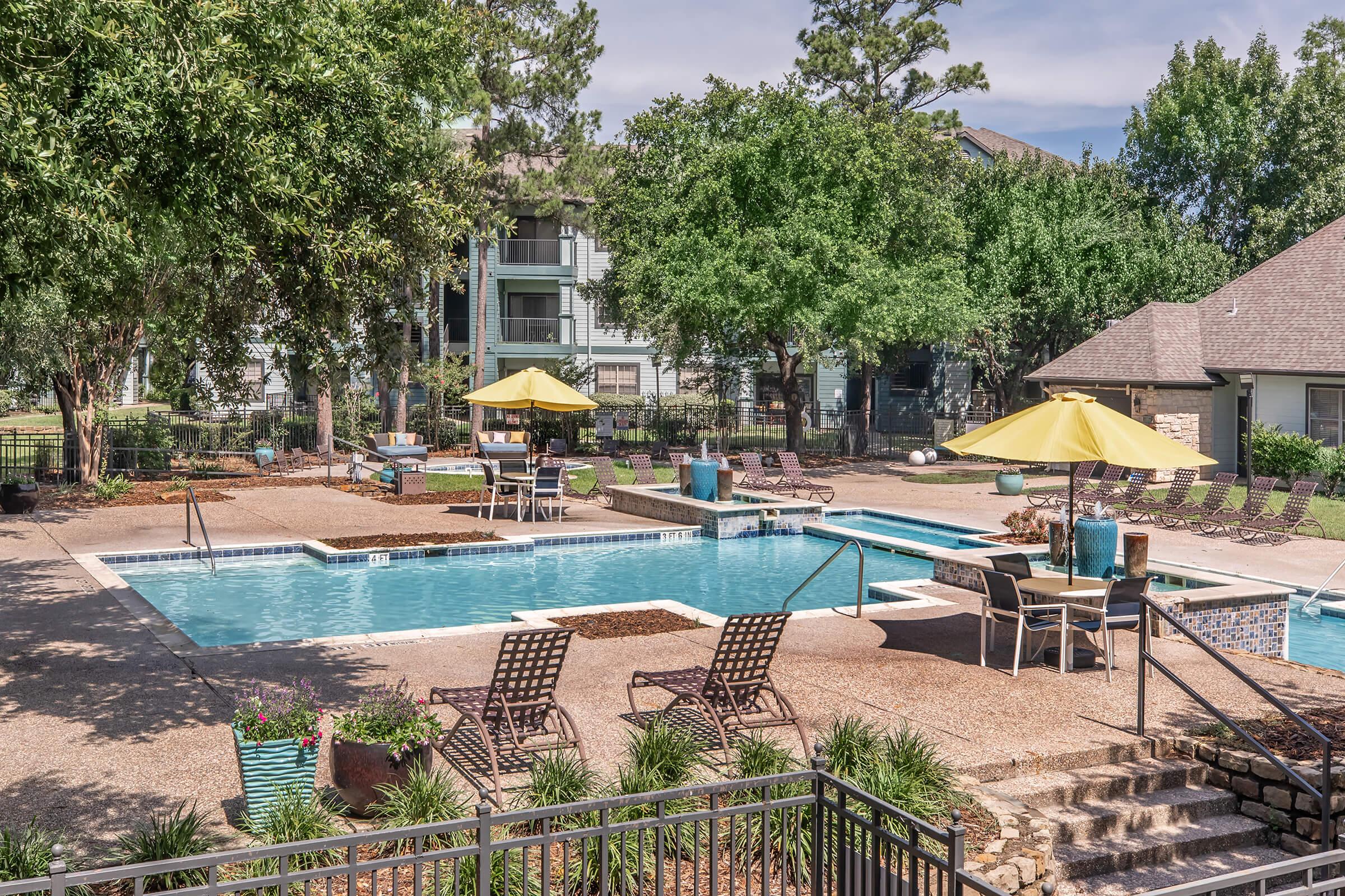 A well-maintained outdoor swimming pool surrounded by lounge chairs and umbrellas. The area features landscaped greenery and walkways, with several trees providing shade. Apartments can be seen in the background, enhancing the community ambiance.