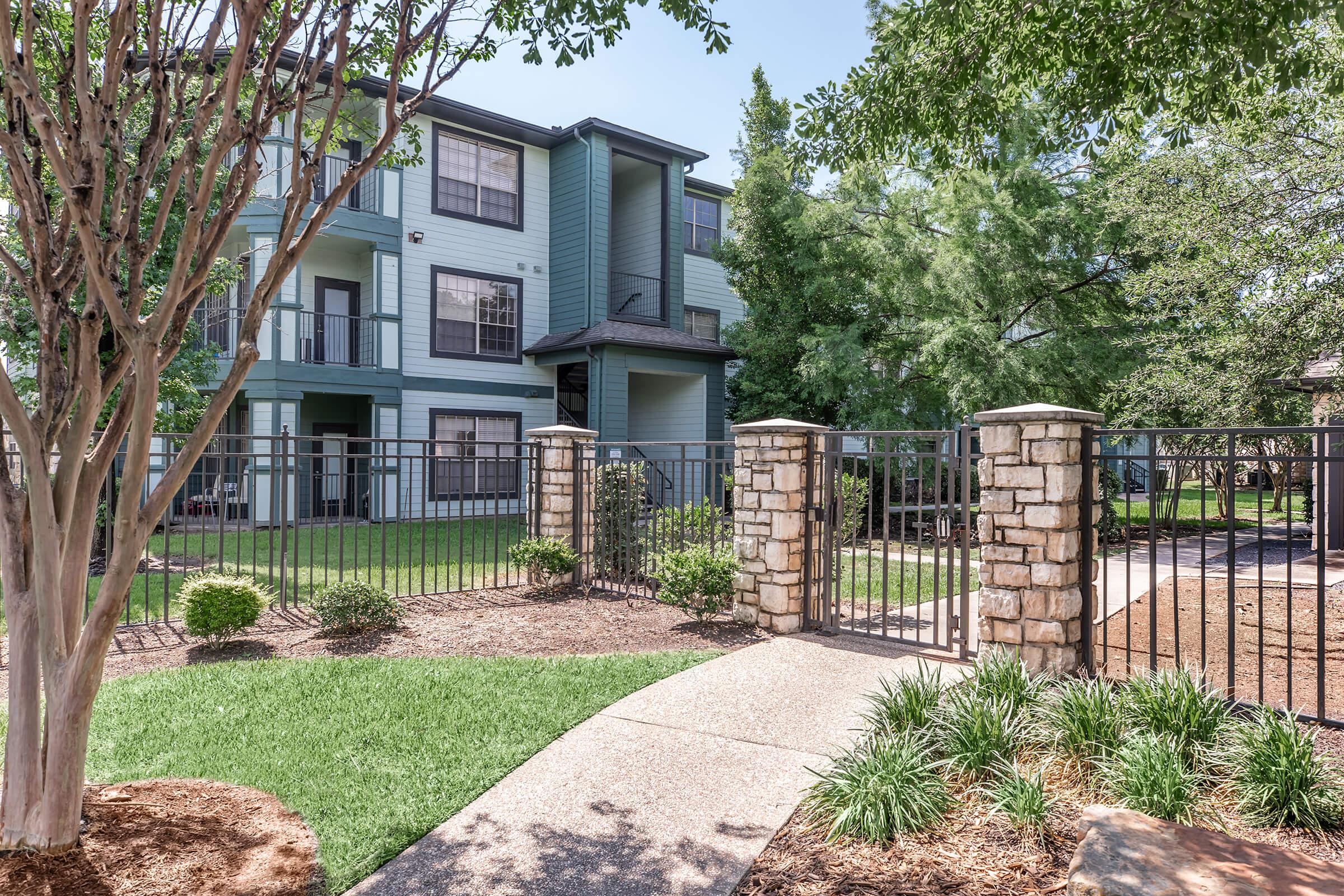 Green grass and landscaped shrubs surround a gated entrance leading to a three-story apartment building. The building features a combination of blue and beige siding, with large windows and balconies. Trees provide shade, creating a peaceful outdoor environment.