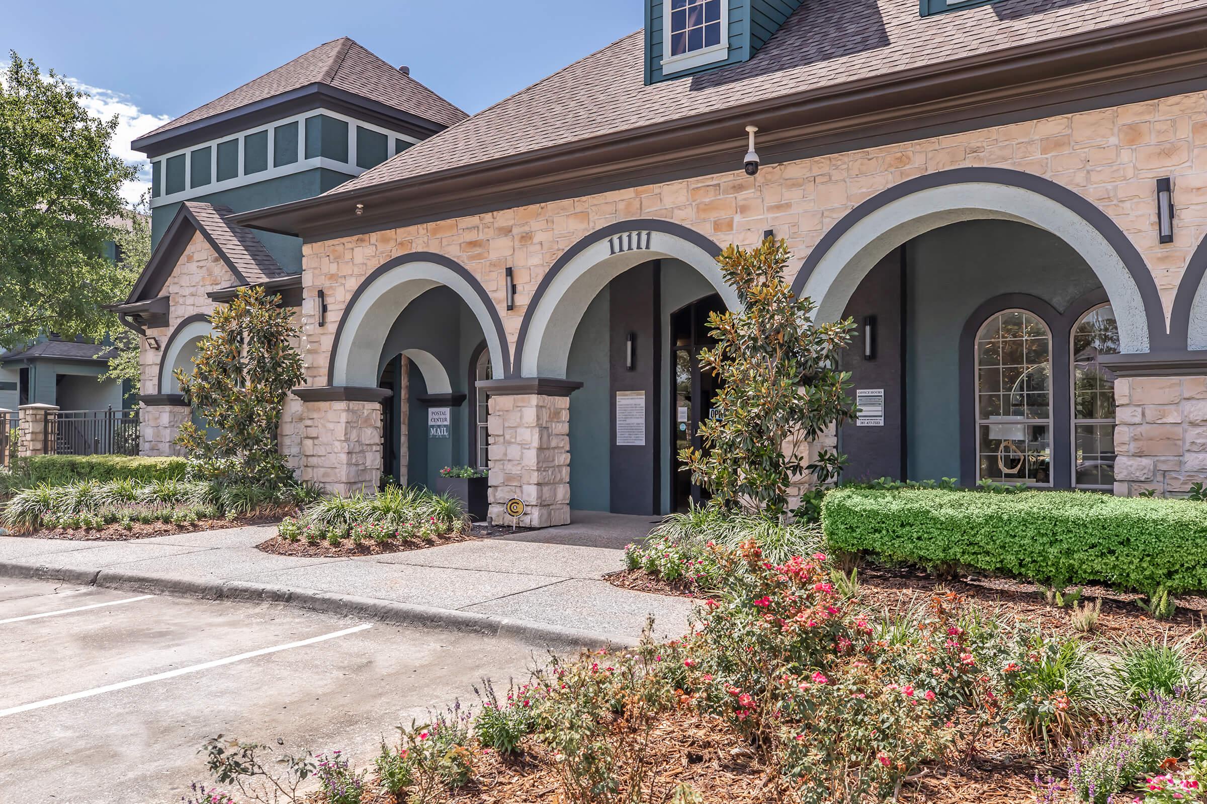 A stone and stucco building featuring arched doorways and large windows, surrounded by landscaped greenery and colorful flowers. The roof has a dark shingle design, and the overall style reflects a mix of modern and classic architectural elements. Parking area in front is visible.