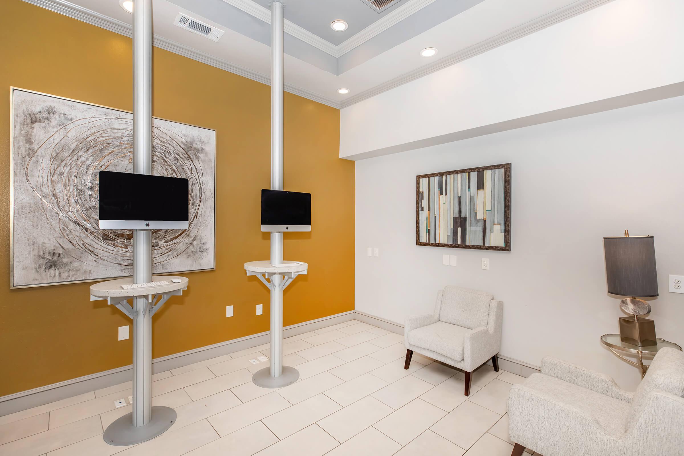 A modern waiting area featuring two wall-mounted computer stations on sleek stands, a framed abstract artwork on the wall, a stylish lamp on a side table, and a contemporary gray armchair on light-colored tiled flooring, complemented by a warm yellow accent wall.