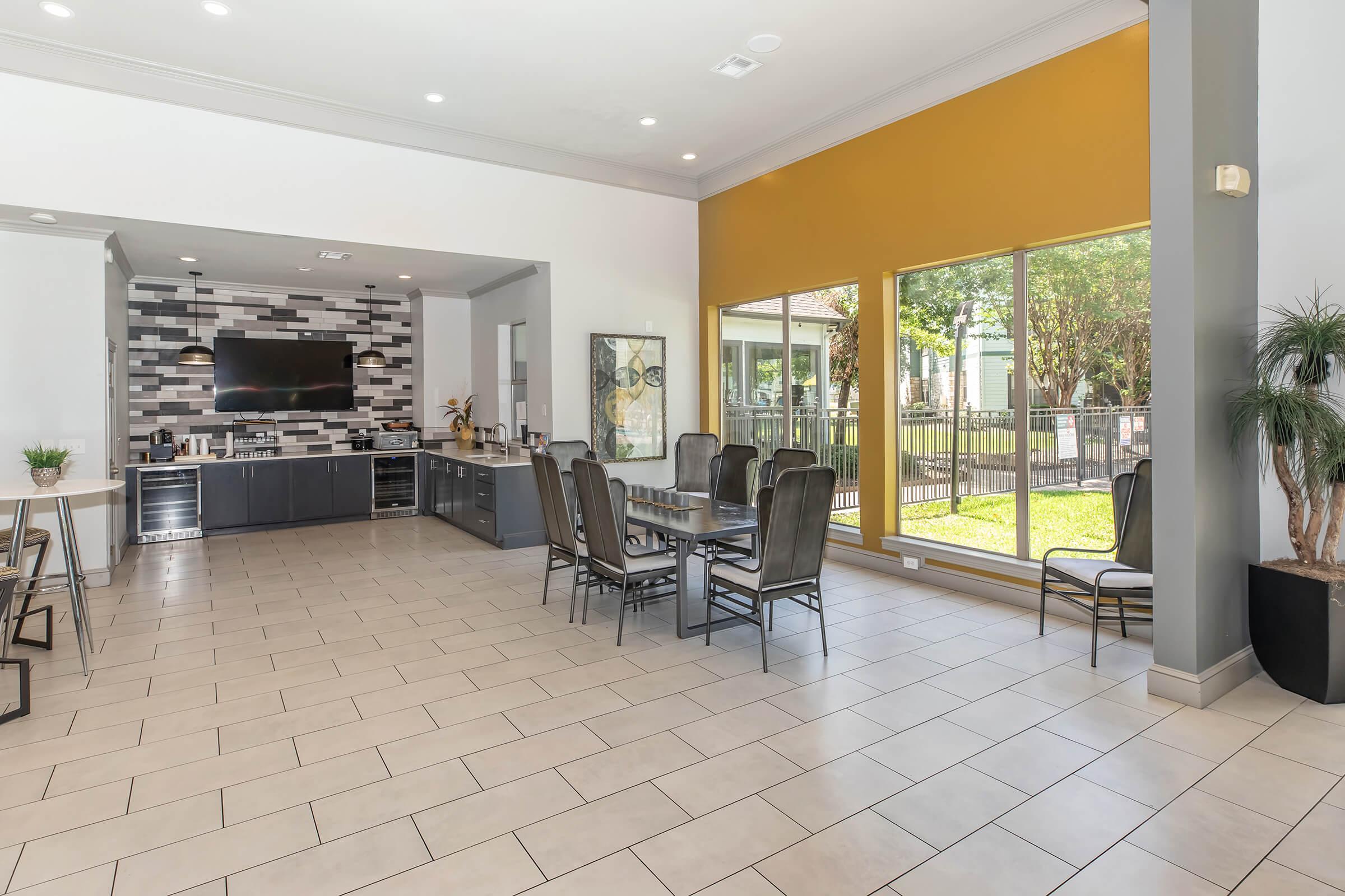 Modern open-concept dining area with large windows offering natural light. Features a stylish wall with a mounted TV, contemporary cabinetry, and a mix of dining furniture. Neutral tile floors complement the bright yellow accent wall and greenery visible outside. Ideal for social gatherings or casual dining.
