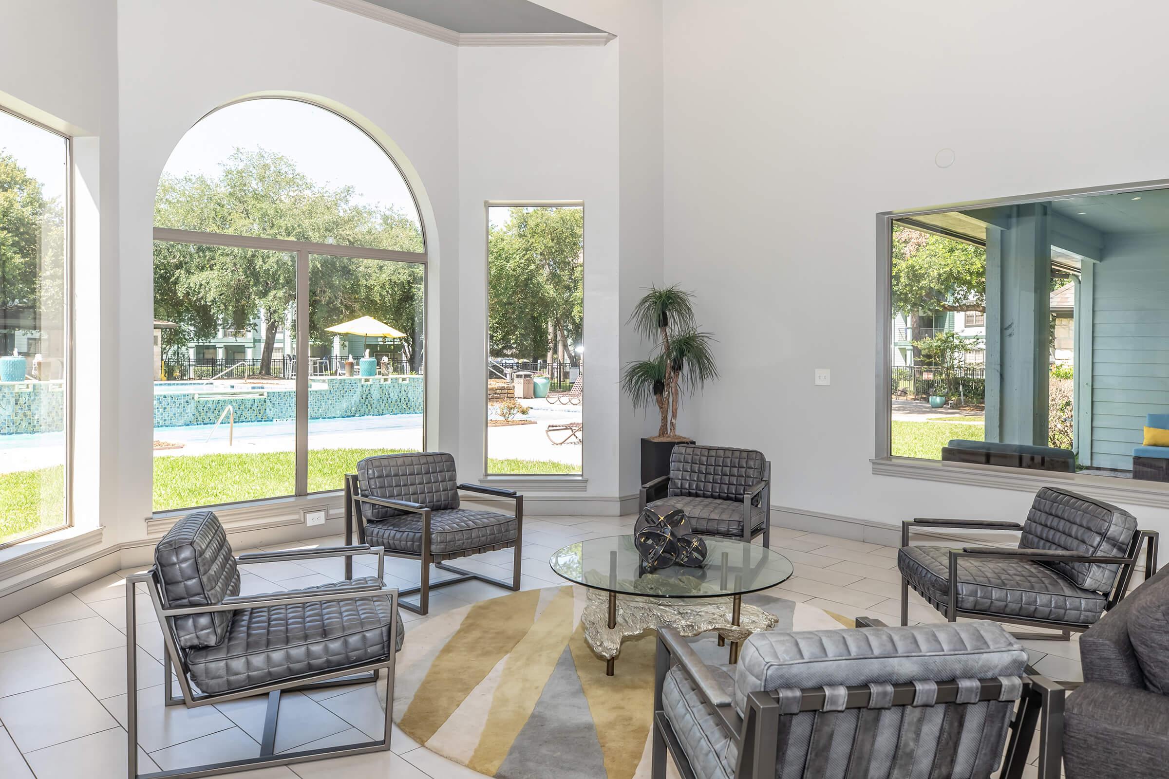 A modern lounge area featuring stylish gray upholstered chairs and a glass coffee table, with large windows showcasing a swimming pool and outdoor seating area visible in the background. The space is bright and airy, decorated with minimalistic furnishings and natural light filtering through.