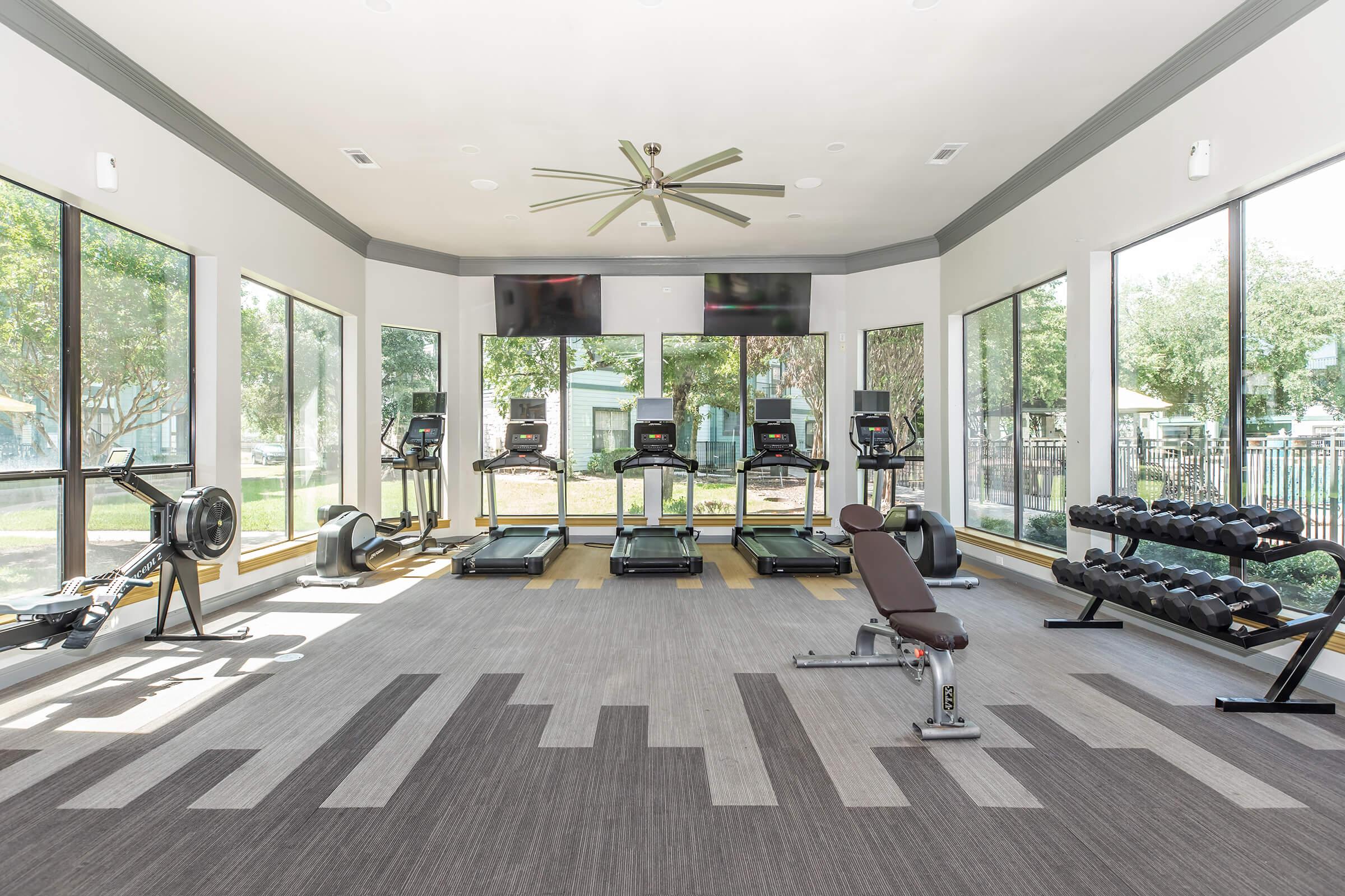 A bright, spacious gym features multiple treadmills, an indoor rowing machine, and a bench with dumbbells. Large windows provide natural light and views of the outdoors, while a ceiling fan circulates air. Two televisions are mounted on the wall for entertainment during workouts.