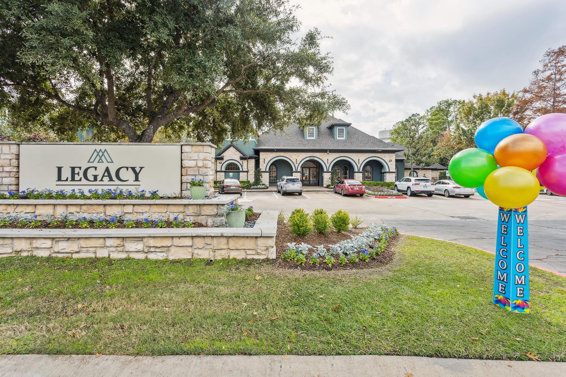 Sign for "Legacy" with landscaped area, featuring colorful balloons and a "Welcome" message on the right. In the background, a building with arches and several parked cars is visible under a partly cloudy sky. The scene includes green grass and flowerbeds.