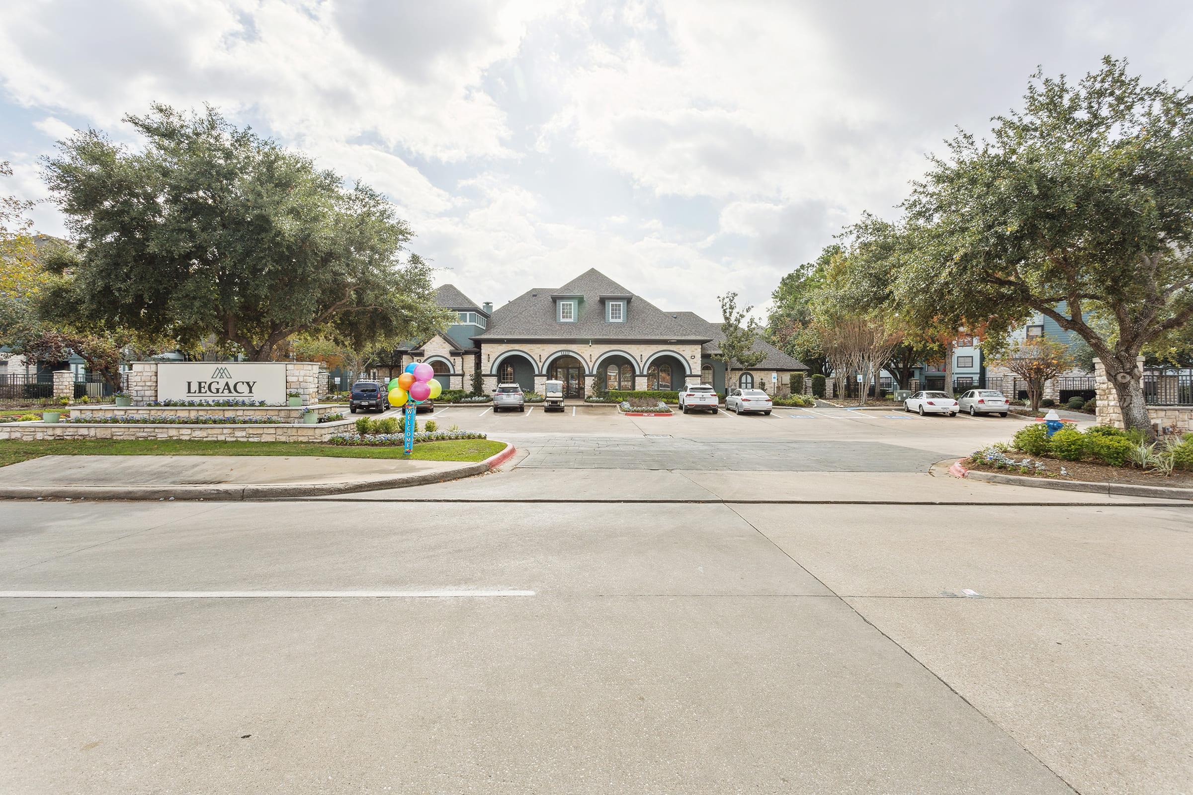 View of the Legacy apartment complex entrance featuring a large sign, colorful balloons at the entrance, and several parked cars. Surrounding the area are trees and landscaped gardens, set against a backdrop of cloudy skies.