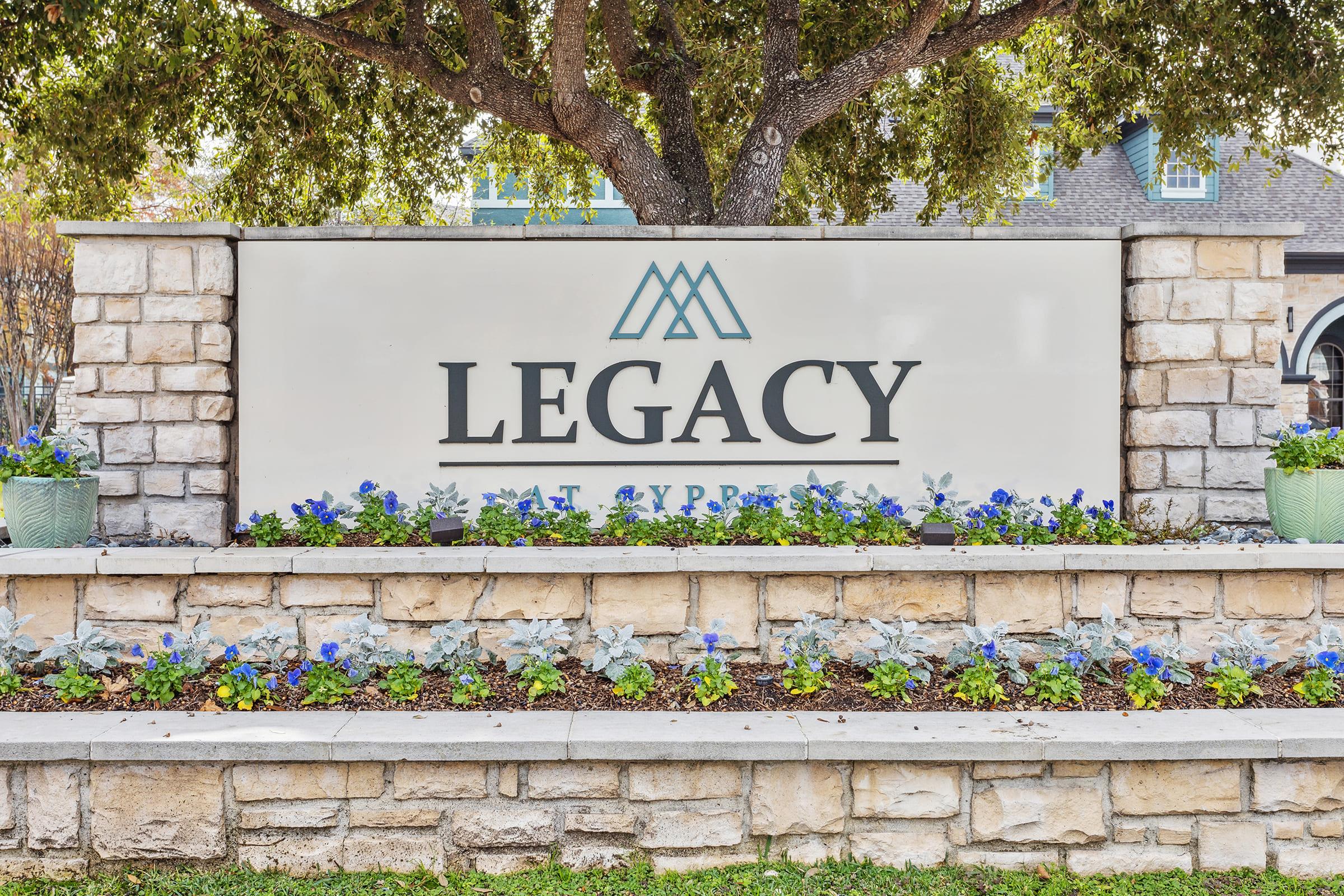 Sign for "Legacy" at Cypress, featuring a modern design with stone elements. The sign is surrounded by colorful flowers and greenery, set beneath a large tree, creating a welcoming entrance to the property.