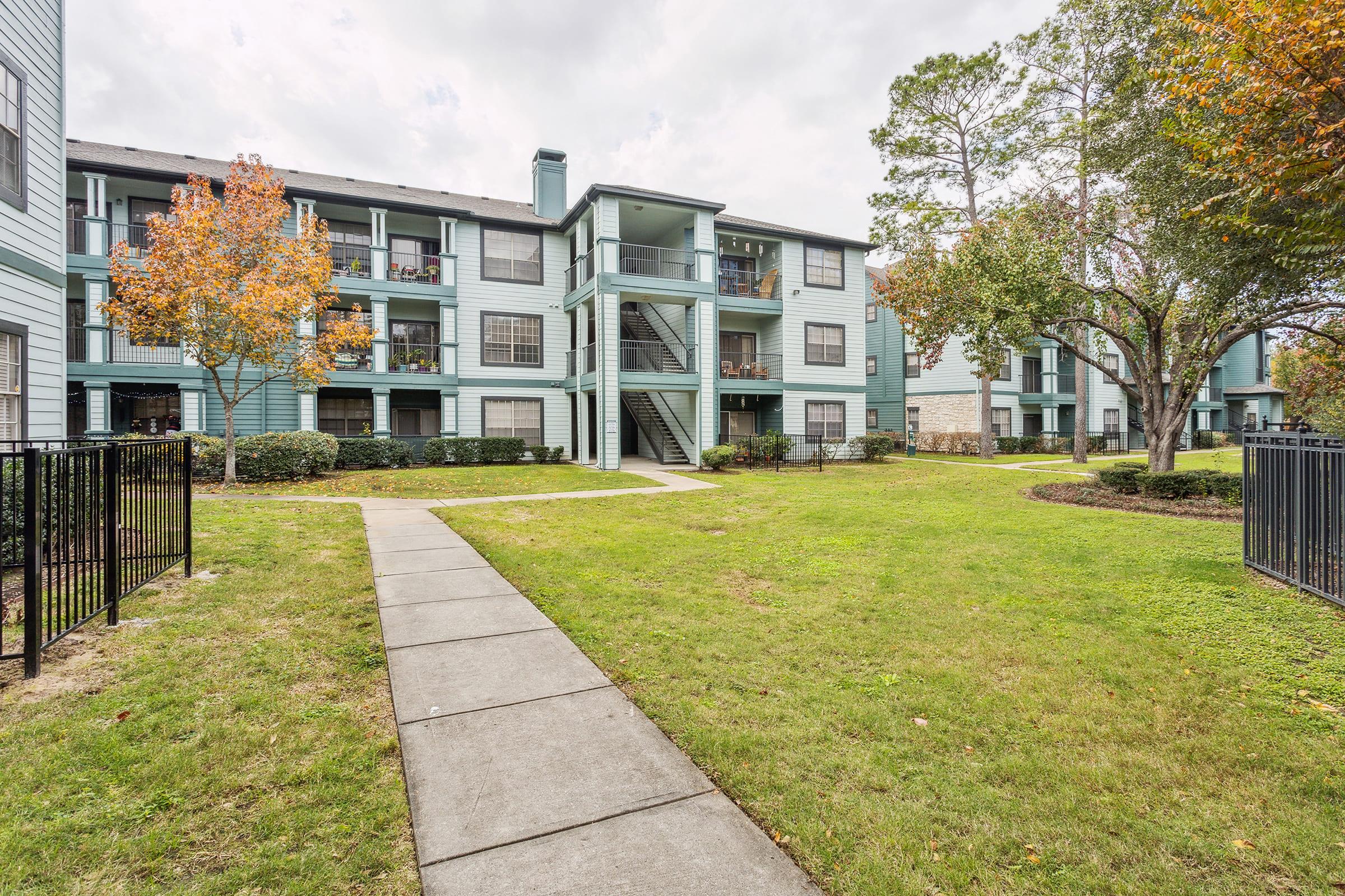 View of a multi-story apartment complex with light blue siding, featuring balconies and surrounded by greenery. A paved walkway leads through a manicured lawn, with trees showing autumn foliage. The setting has a peaceful, residential atmosphere.