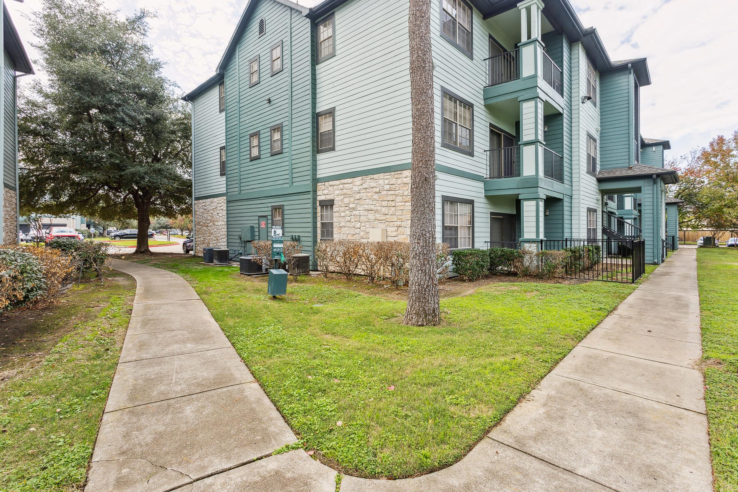 A residential apartment complex featuring a green exterior with multiple floors. The image shows two pathways diverging in front of the building, surrounded by well-maintained grass and shrubs. Trees are visible in the landscape, contributing to a pleasant outdoor environment.