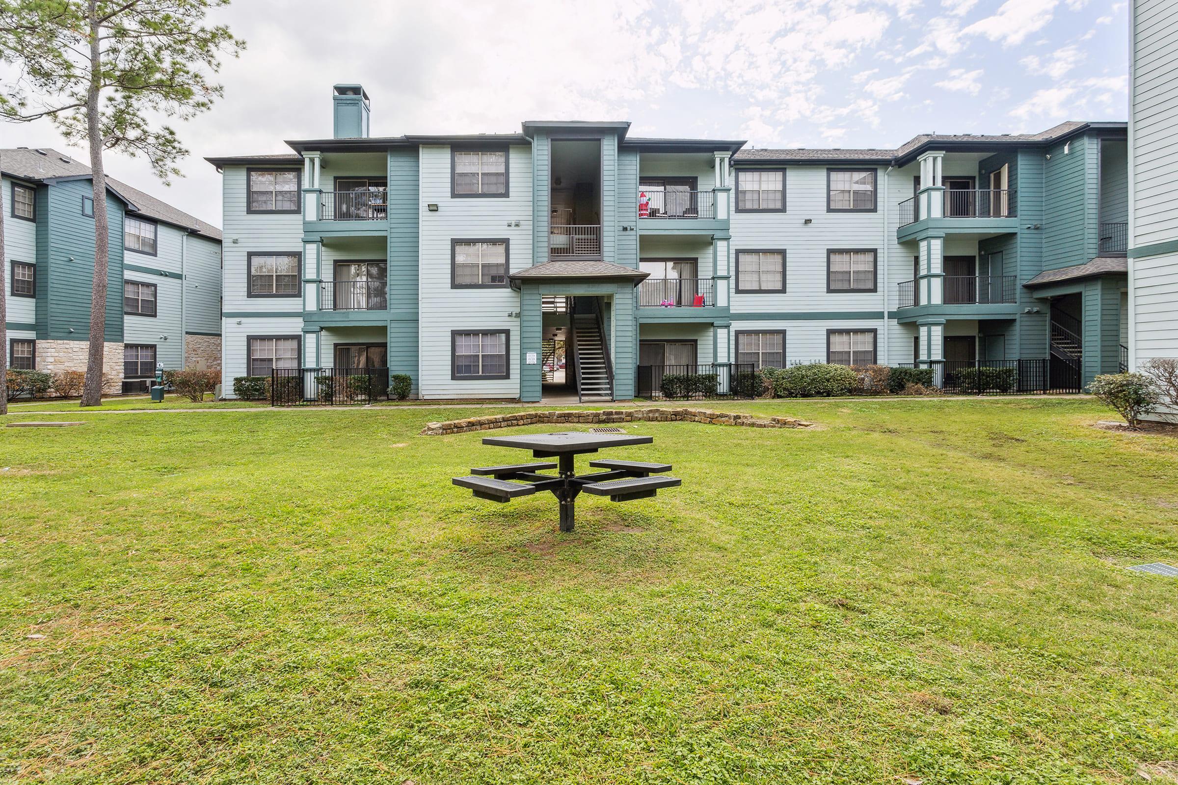 A multi-story apartment building with a light blue exterior, featuring balconies and a central grassy area. In the foreground, there is a black picnic table placed on the lawn. Trees and shrubs surround the building, under a cloudy sky.