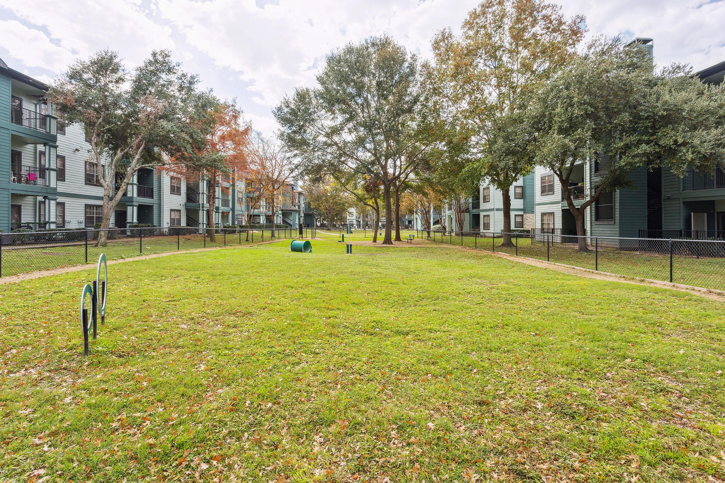 Open green grassy area surrounded by residential apartment buildings. Trees with fall foliage are visible, and there is a fenced boundary. A small dog play area can be seen, along with a few walking paths. The sky is partly cloudy, creating a serene atmosphere.