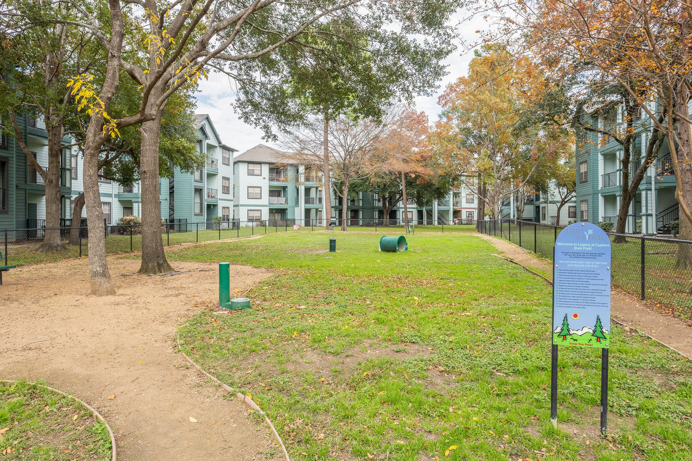 A spacious green area in an apartment complex featuring trees and well-maintained lawn. There are walking paths, a small pet waste station, and a sign with information about the area. Surrounding the space are multi-story apartment buildings, with a mix of autumn foliage on the trees.