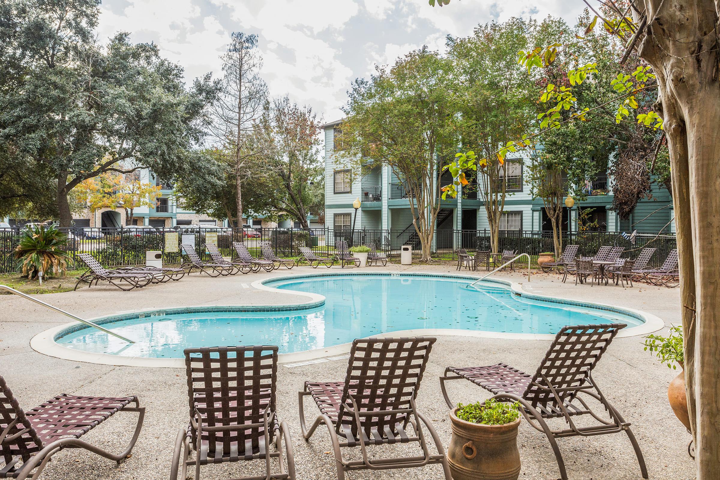 A clean, inviting pool area featuring a heart-shaped swimming pool surrounded by lounge chairs. Lush greenery surrounds the space, with trees and plants enhancing the outdoor ambiance. Nearby, multiple buildings offer residential views, creating a relaxing atmosphere for residents and guests.