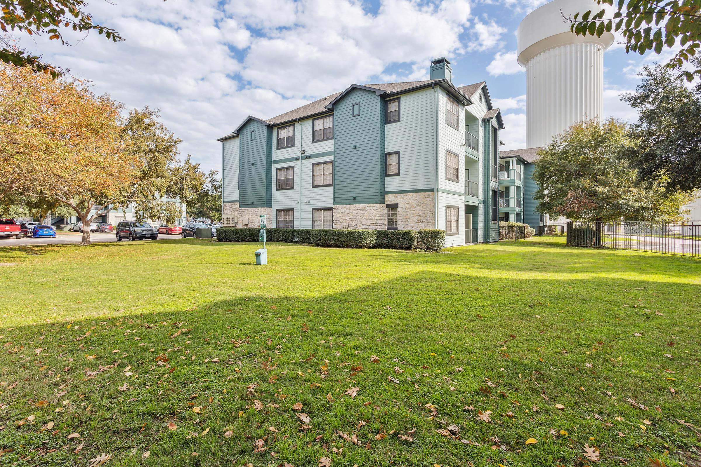 A two-story green apartment building with stone accents, surrounded by a grassy lawn and trees. In the background, there is a tall water tower. Several parked cars are visible, and the sky is partly cloudy with scattered clouds.