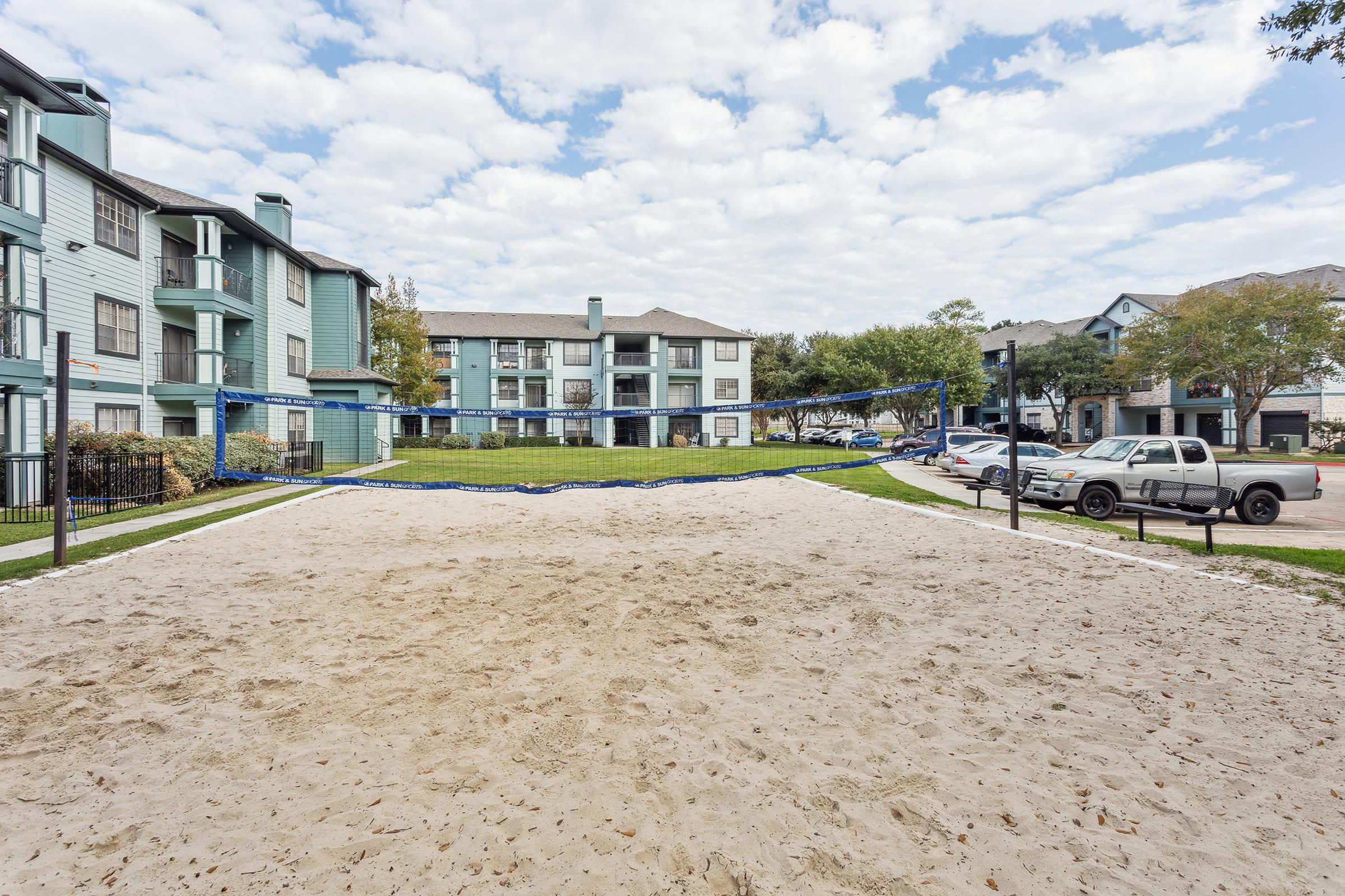 Sand volleyball court with a blue net set up in a grassy area surrounded by apartment buildings. In the background, several parked cars are visible, and the sky is partly cloudy. The setting is inviting and suitable for recreational activities.