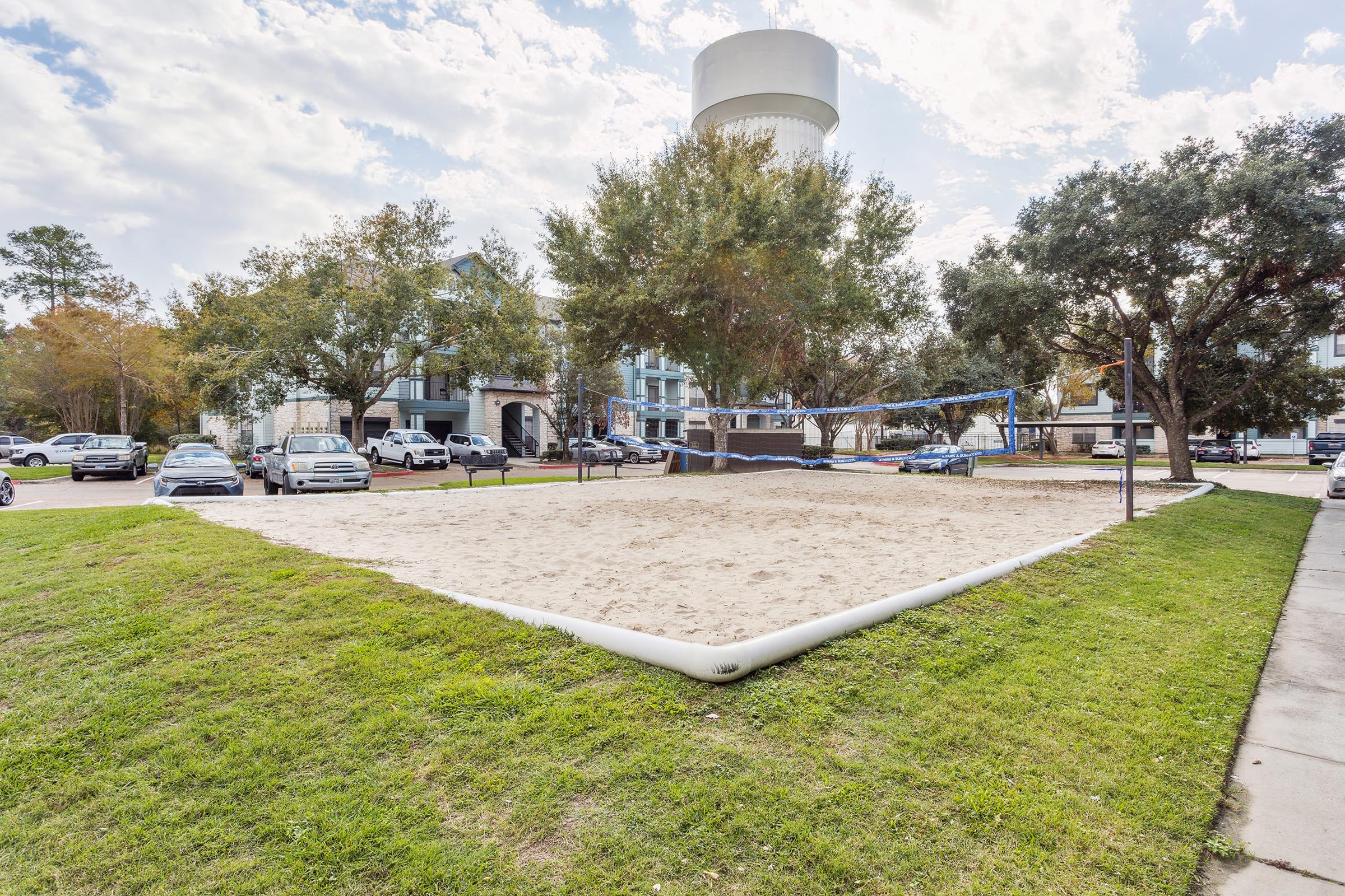A sandy volleyball court surrounded by grass and trees, with a water tower visible in the background. Several parked cars are lined up along the street, and an apartment building can be seen nearby under a partly cloudy sky.