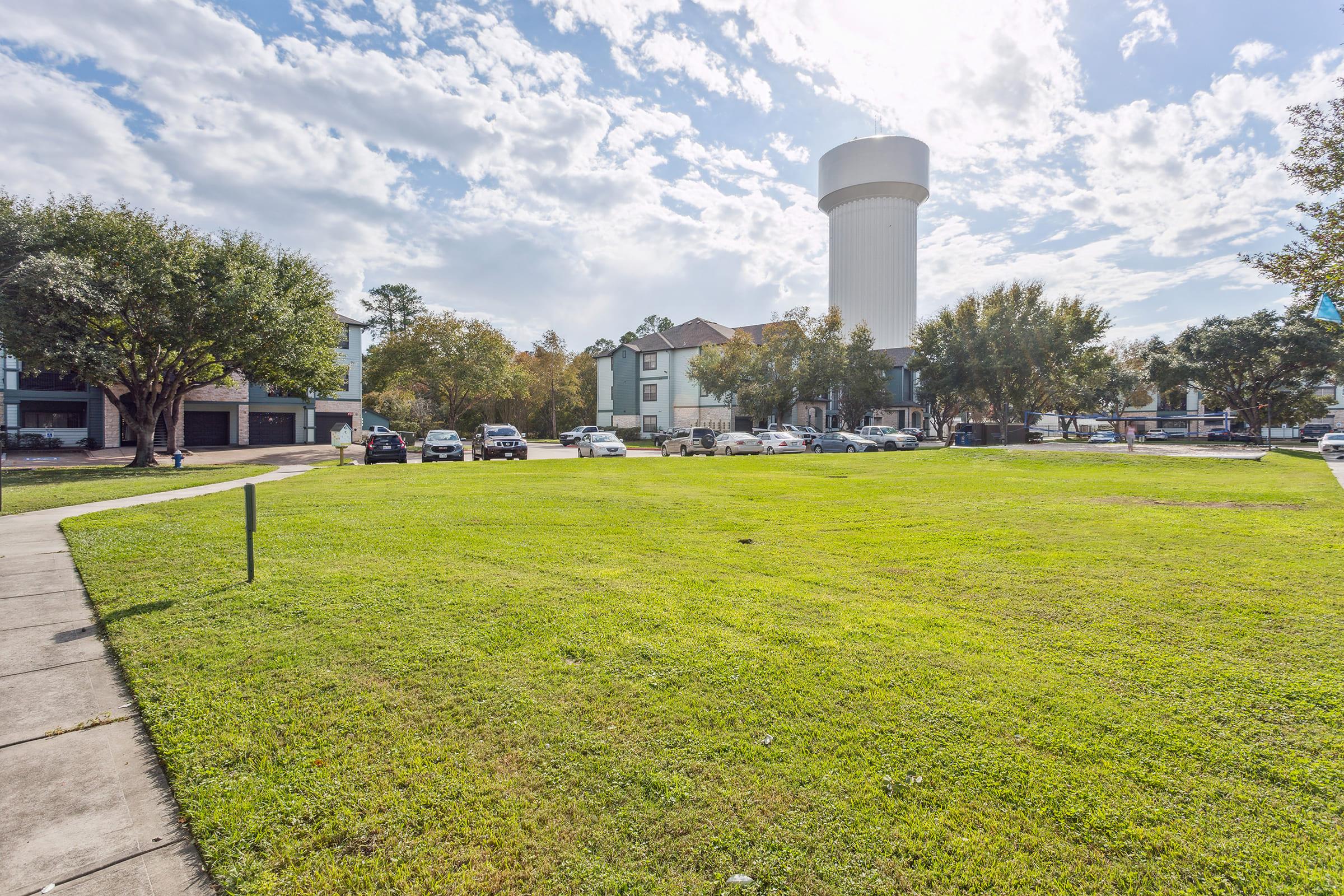 A spacious green lawn is surrounded by several parked cars and residential buildings. In the background, a tall, white water tower stands against a partly cloudy sky. The scene is bright and inviting, showcasing a pleasant neighborhood setting.
