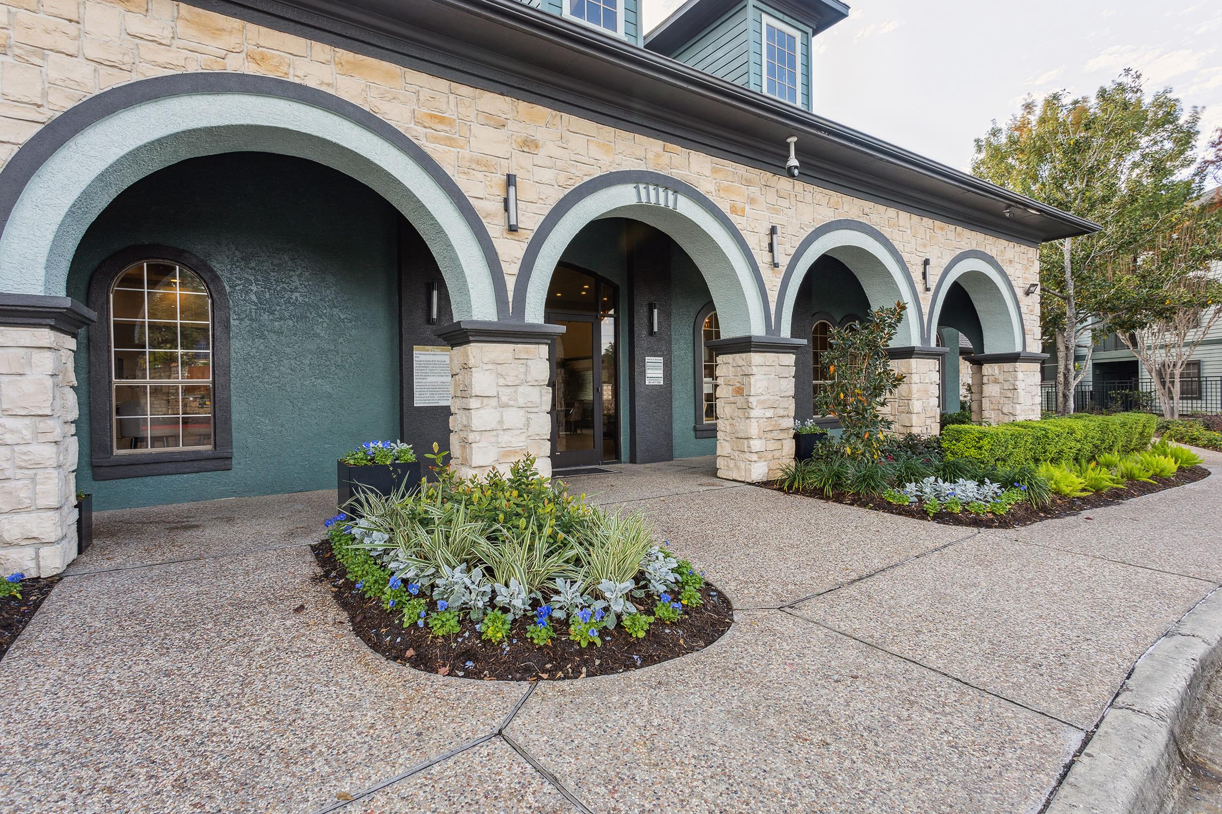 Exterior view of a building featuring stone arches and a landscaped entryway with various plants and flowers. The pathway is made of textured stone, leading to double doors framed by large windows. The overall architecture combines modern design with natural elements.