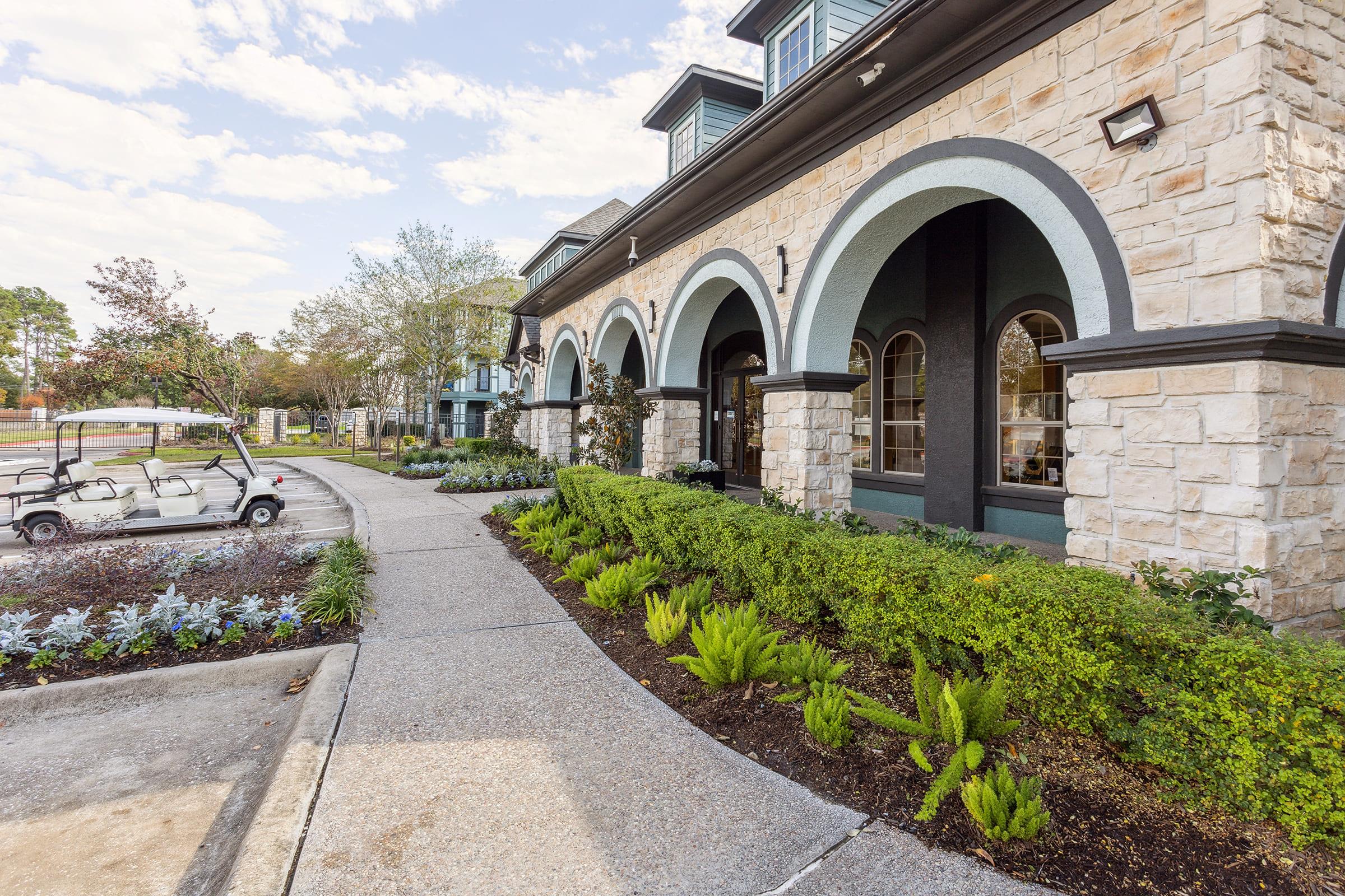 A landscaped pathway leads to the entrance of a modern building with stone arches. Colorful plants and shrubs line the walkway, adding greenery. A golf cart is visible parked nearby, with trees and the sky in the background, indicating a well-maintained outdoor area.