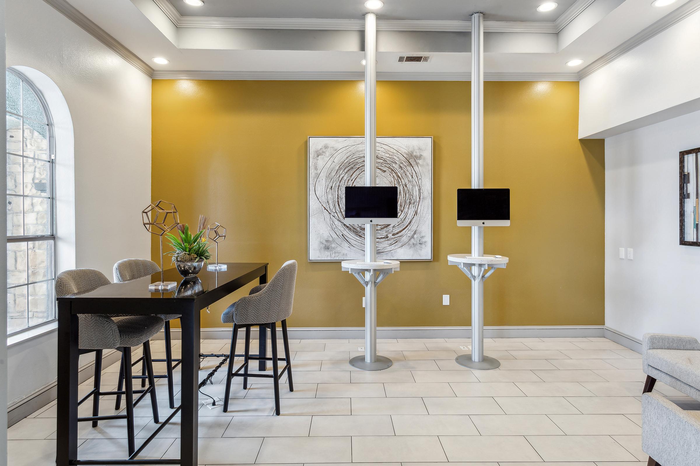 A modern lobby area featuring a yellow accent wall, two freestanding monitor stands, a dark table with high-backed chairs, and a decorative plant. Large windows allow natural light, creating a welcoming atmosphere suitable for casual meetings or waiting areas.