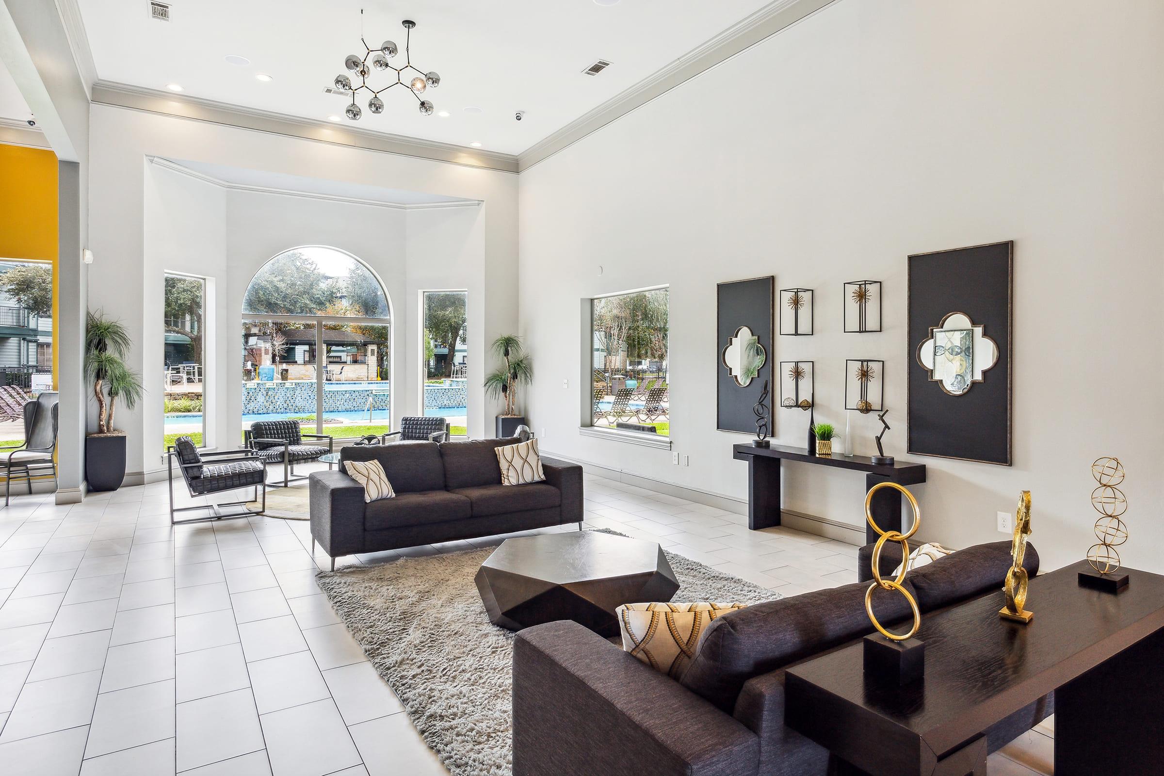 A modern, stylish lobby featuring light-colored walls and tiled flooring. The space includes two sofas, a coffee table, and decorative elements like sculptures and wall art. Large windows offer a view of a pool area outside, letting in natural light and creating a welcoming atmosphere.