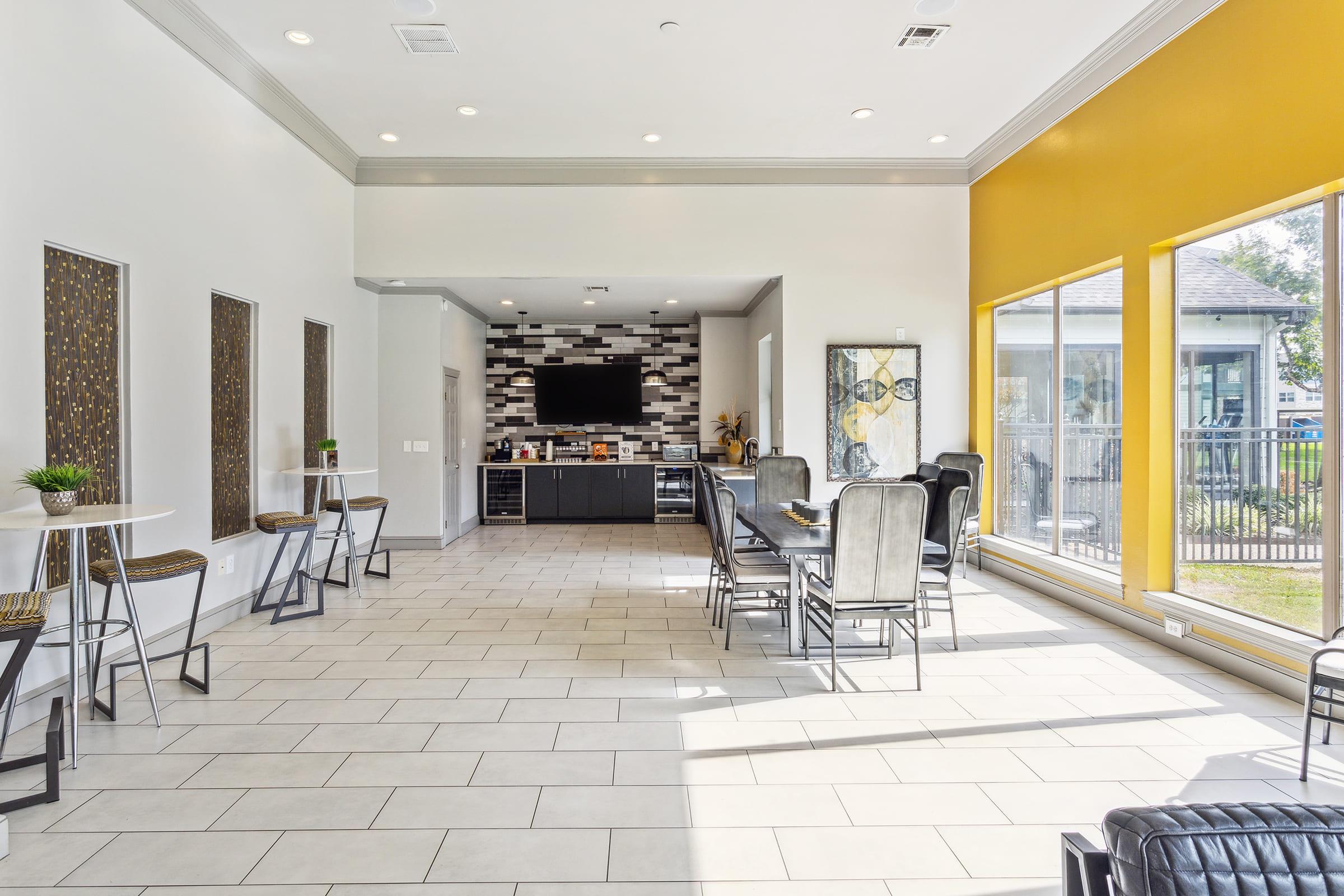 A modern, well-lit common area featuring several tables and chairs. There are large windows with yellow accents, sleek tile flooring, and contemporary wall art. A black cabinet with a TV and decorative elements is present at the far end, creating an inviting space for socializing or gatherings.