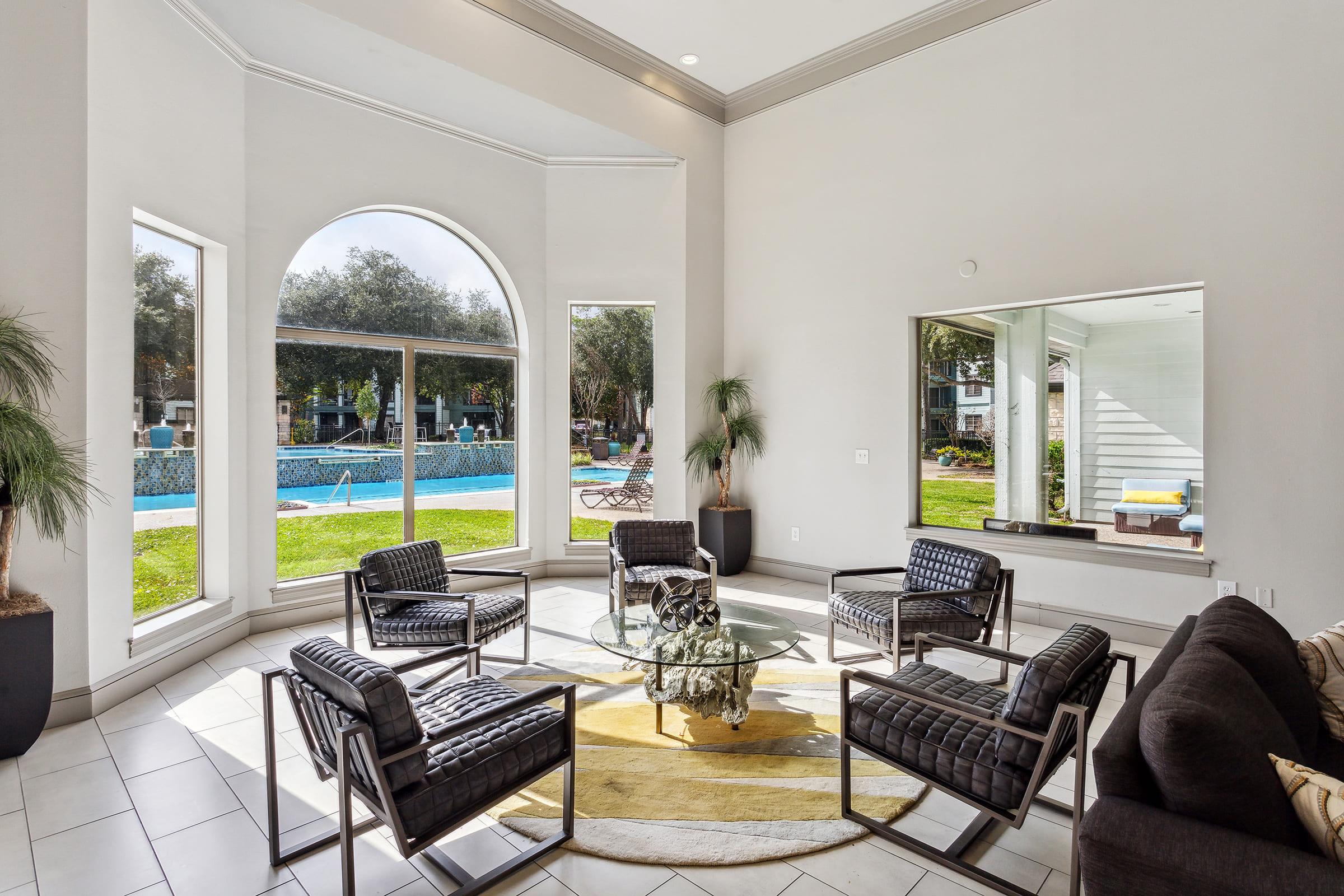 A modern lounge area featuring stylish black chairs and a glass table. Large windows provide a view of a swimming pool and landscaped outdoor space. Natural light illuminates the room, enhancing the inviting atmosphere with potted plants in the corners.
