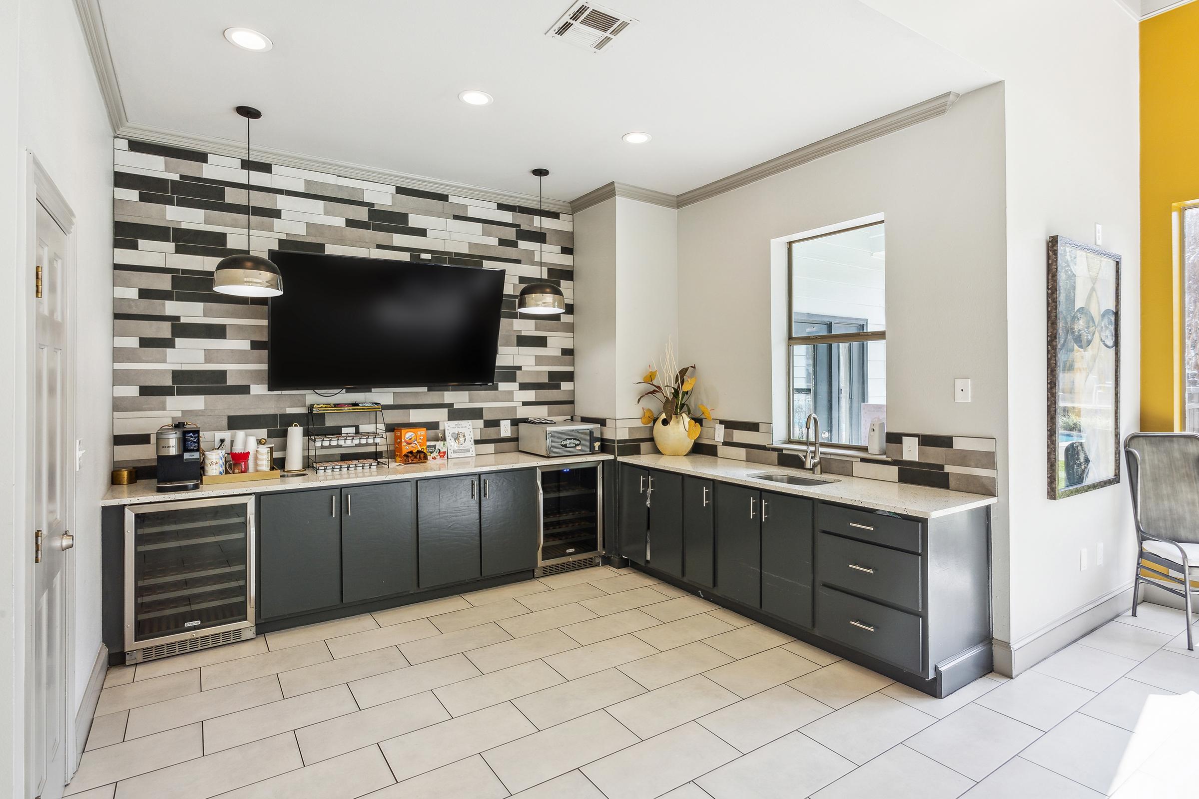 Modern kitchen area featuring a sleek design with gray tiled backsplash, large flat-screen TV mounted on the wall, and a combination of dark cabinets and light countertops. The space includes coffee machines, under-counter refrigerators, and decorative plants, creating a stylish and functional environment.