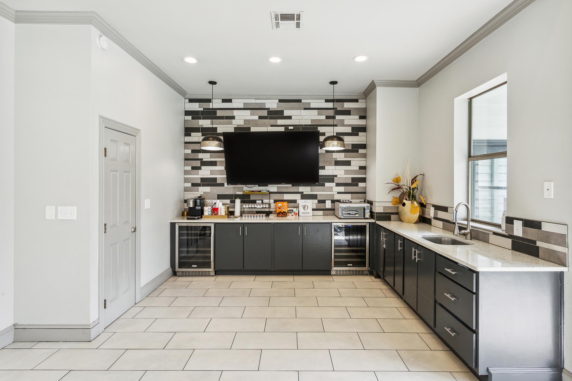 Modern kitchen featuring a sleek gray and white color scheme. It includes a wall-mounted TV, a large countertop with a sink, and dark cabinetry. The backsplash consists of a patterned tile design, and the space is well-lit with overhead lighting. Cabinets show appliances and decorative items.