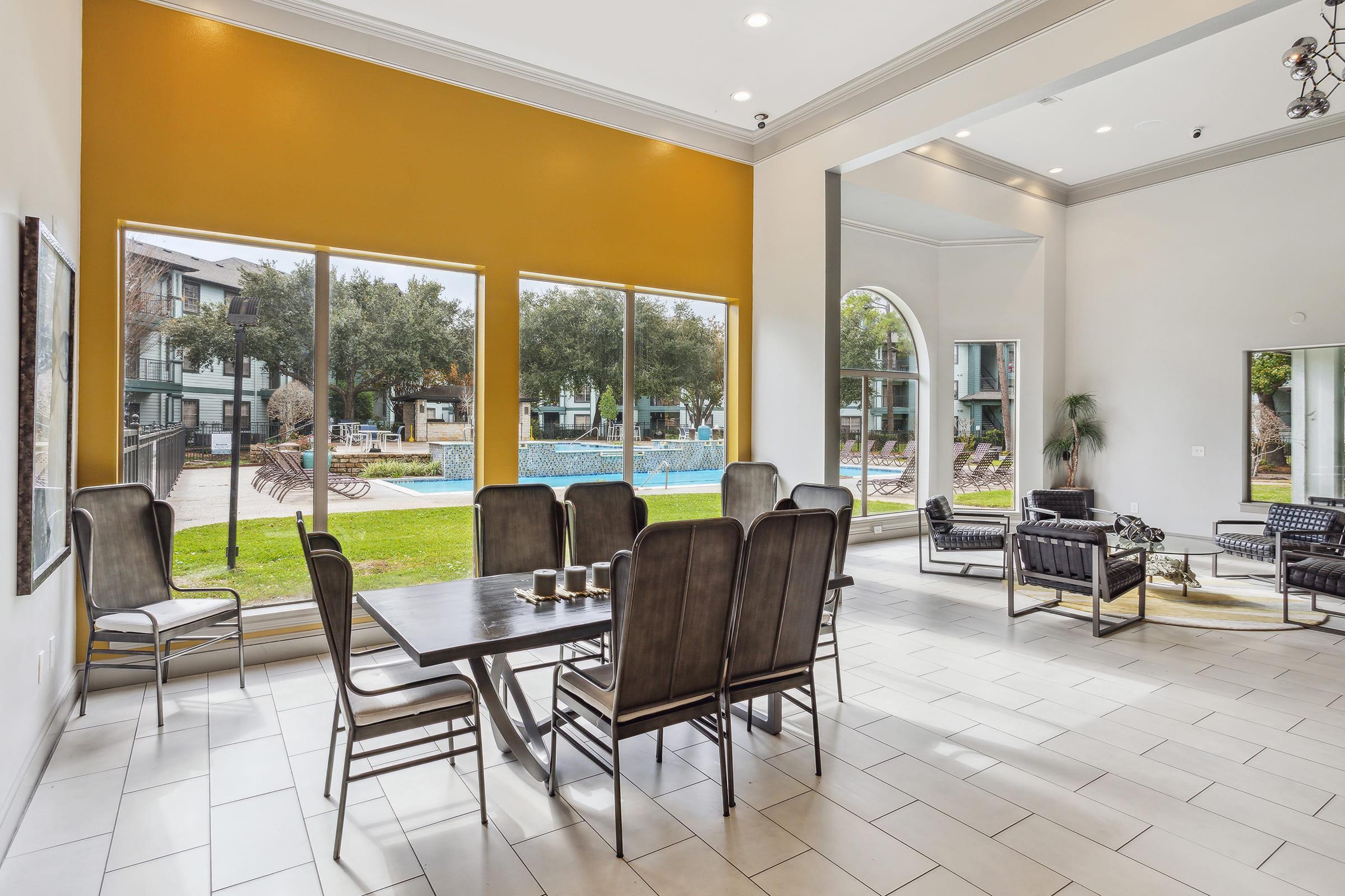 Bright and spacious common area with large windows overlooking a pool. The room features a modern dining table surrounded by chairs, stylish furniture, and a warm yellow accent wall, enhancing the inviting atmosphere. Sunlight floods in, creating a welcoming environment for residents.