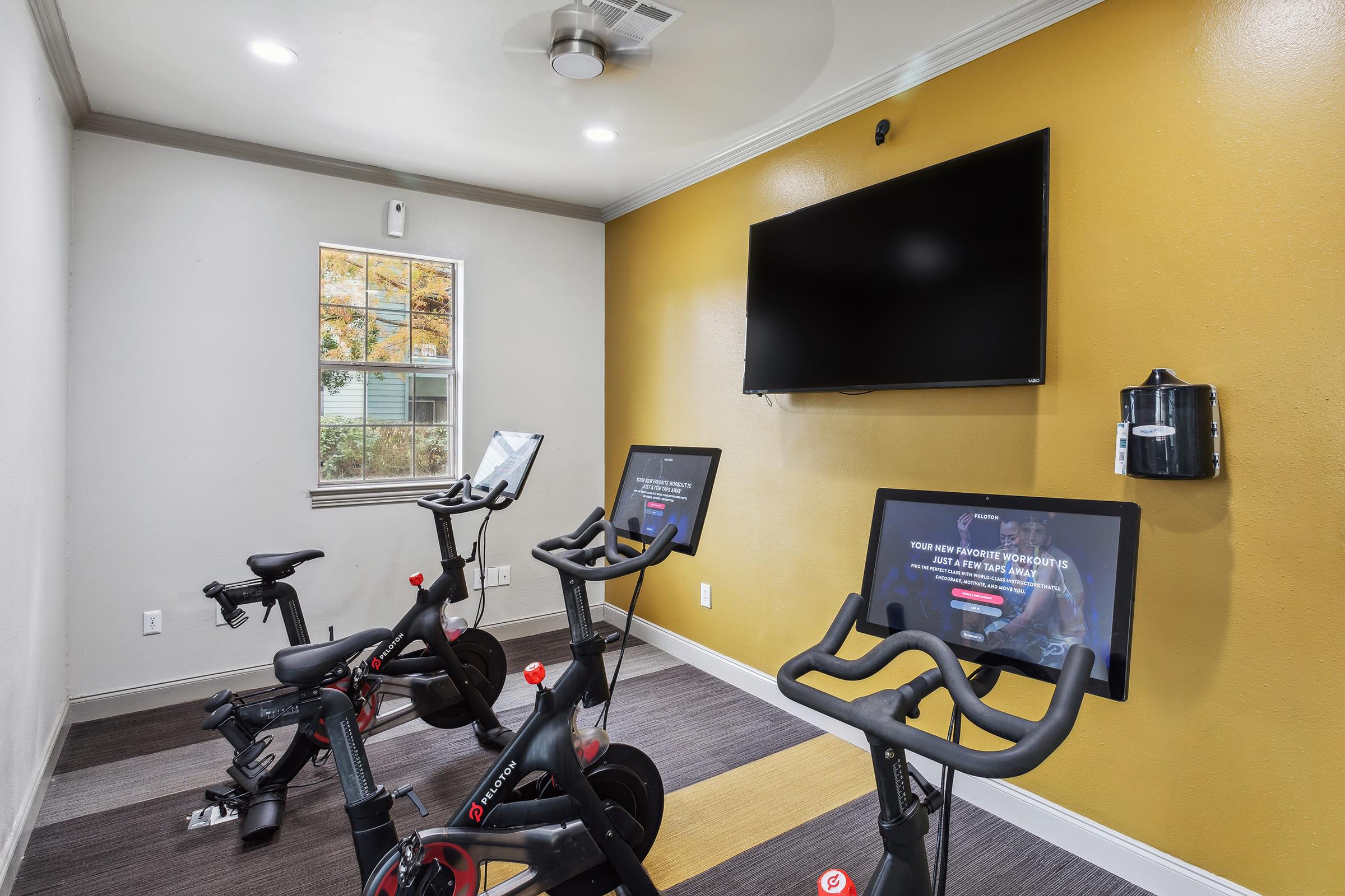 A modern fitness room featuring three stationary bikes with screens displaying workout programs. The walls are painted a warm yellow, and there's a TV mounted on the wall above. A small window allows natural light to enter, enhancing the inviting atmosphere.