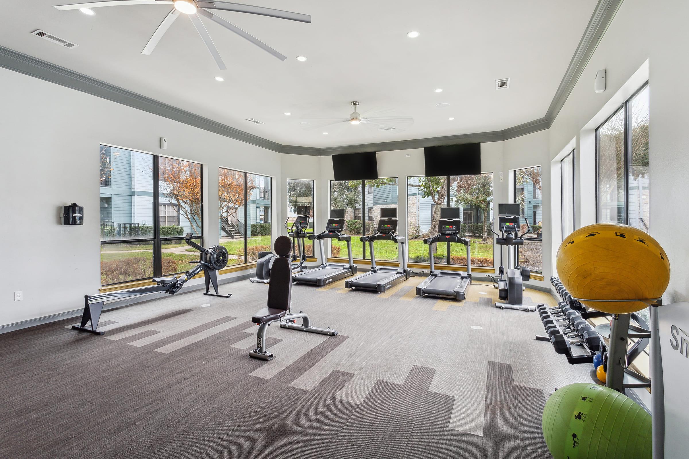 A bright and spacious fitness room featuring several treadmills and exercise bikes, a weight bench, and a selection of weights. Large windows let in natural light and provide views of the outdoor area. The room is well-equipped for workouts with modern amenities and a ceiling fan.