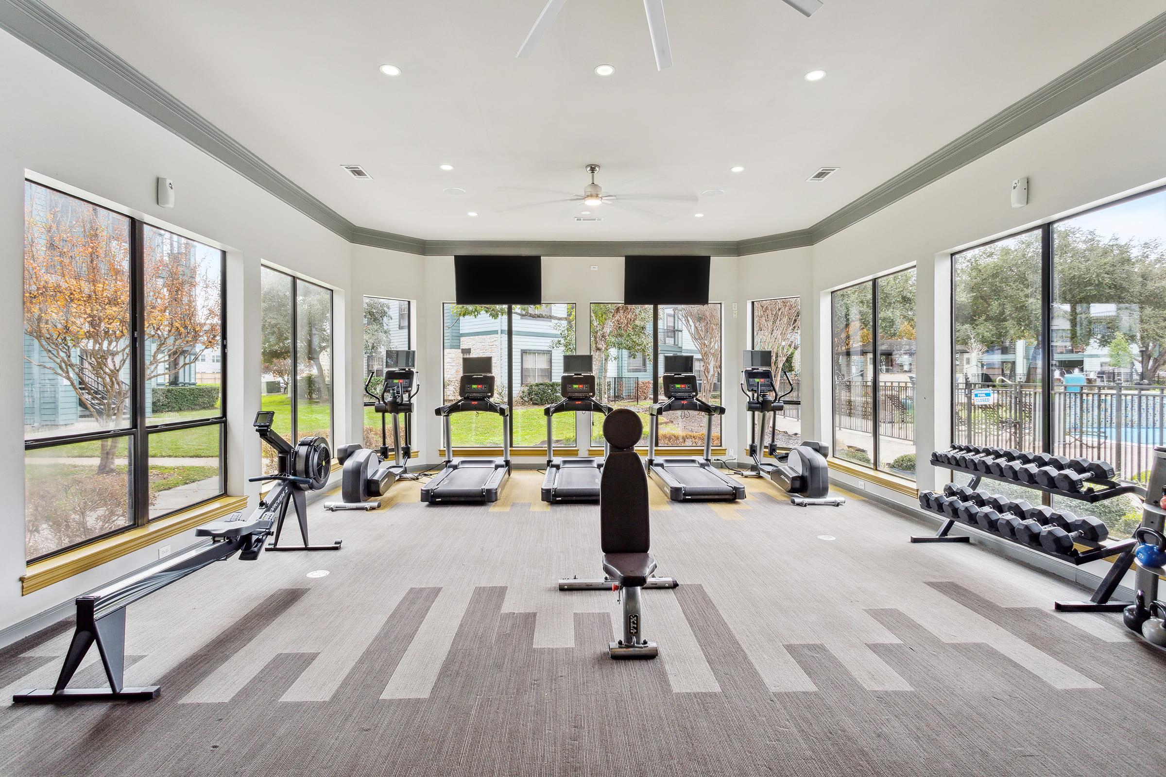 A bright and modern gym with large windows, featuring various exercise equipment including treadmills, elliptical machines, and free weights. The space is well-lit, showcasing a clean, welcoming environment with a view of outdoor greenery.