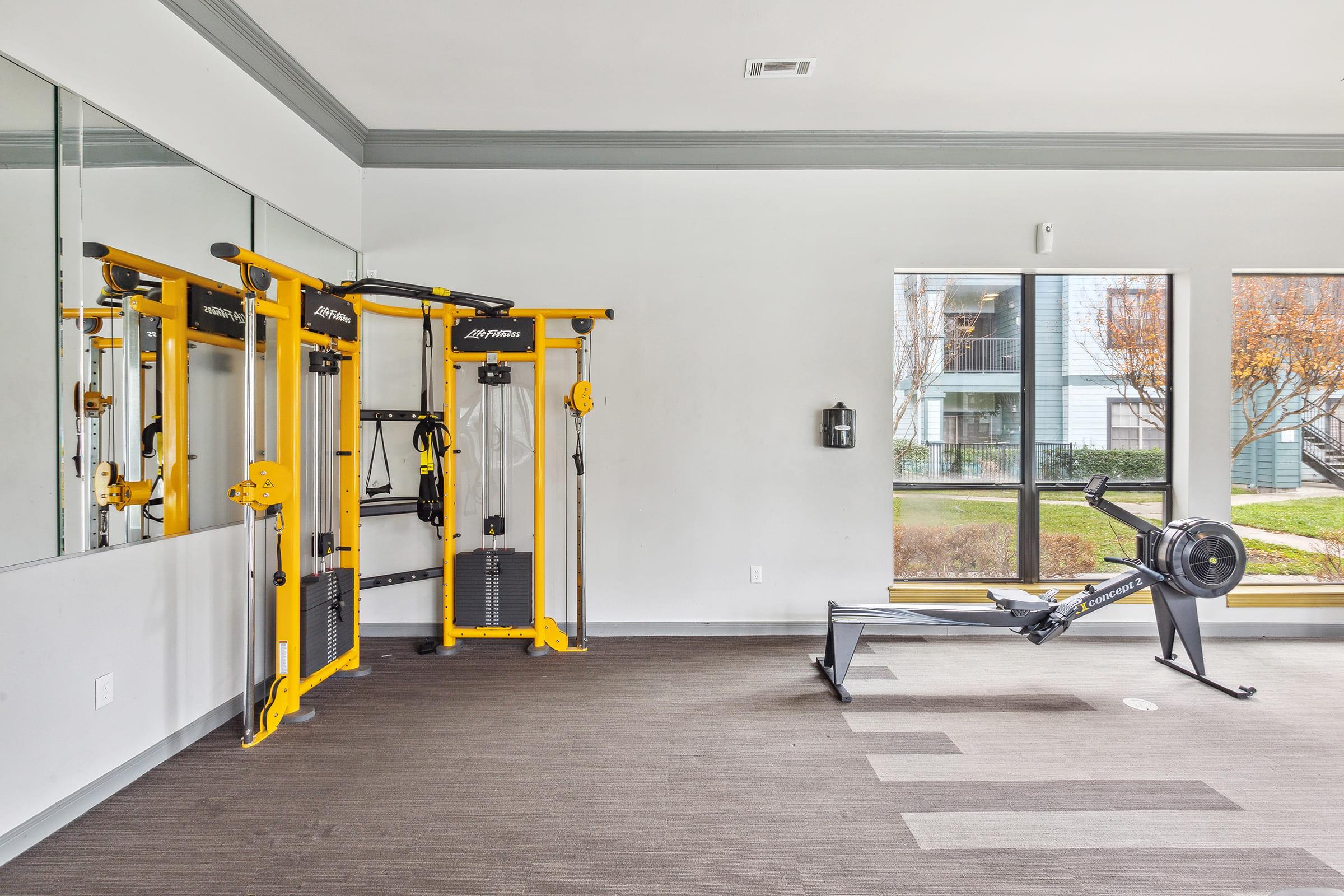 Interior view of a fitness room featuring a yellow multi-gym machine and a rowing machine. The space has large windows providing natural light and a view of the outdoors. The flooring is carpeted in a neutral color, complementing the modern design of the room.