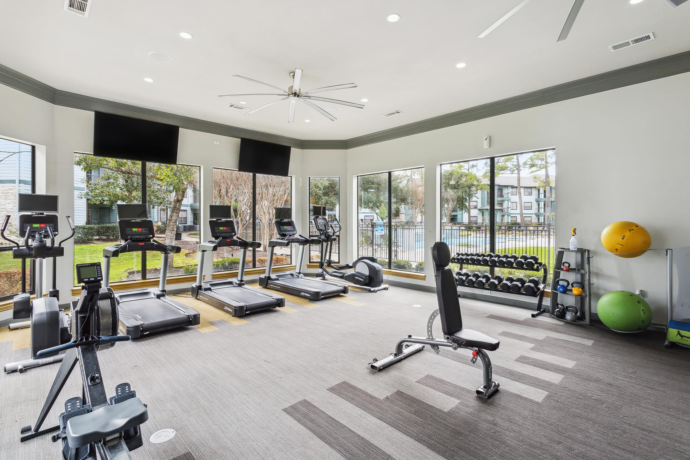 A modern gym interior featuring several treadmills, a weight bench, and free weights. Large windows provide natural light and a view of the outdoors. The space is well-lit and equipped with fitness equipment to accommodate various workout routines.