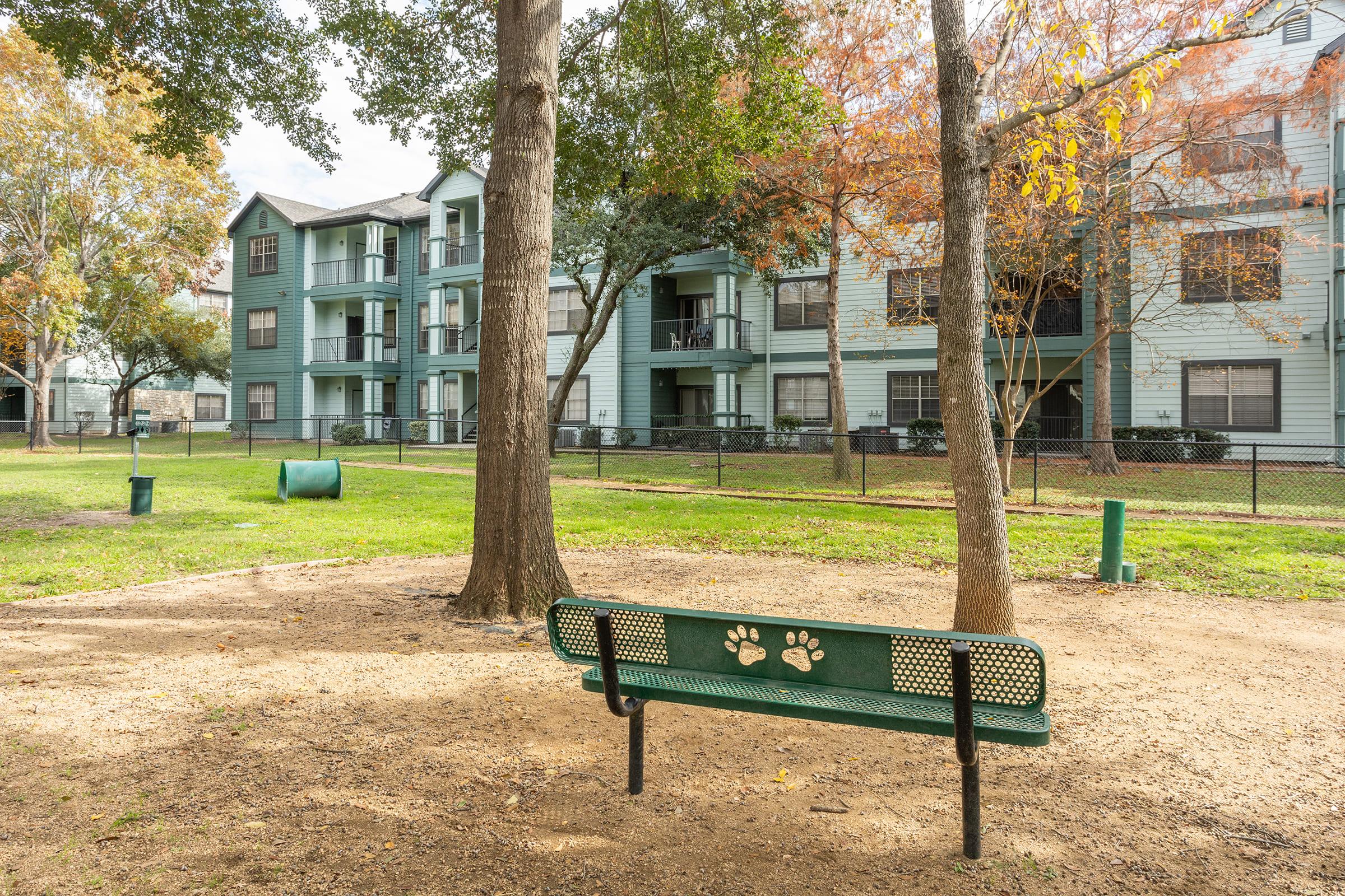 A green bench with paw print designs is situated in a grassy area surrounded by trees, facing a residential apartment complex. The scene features a dirt path, a waste bin for pets, and autumn foliage, creating a peaceful outdoor environment.