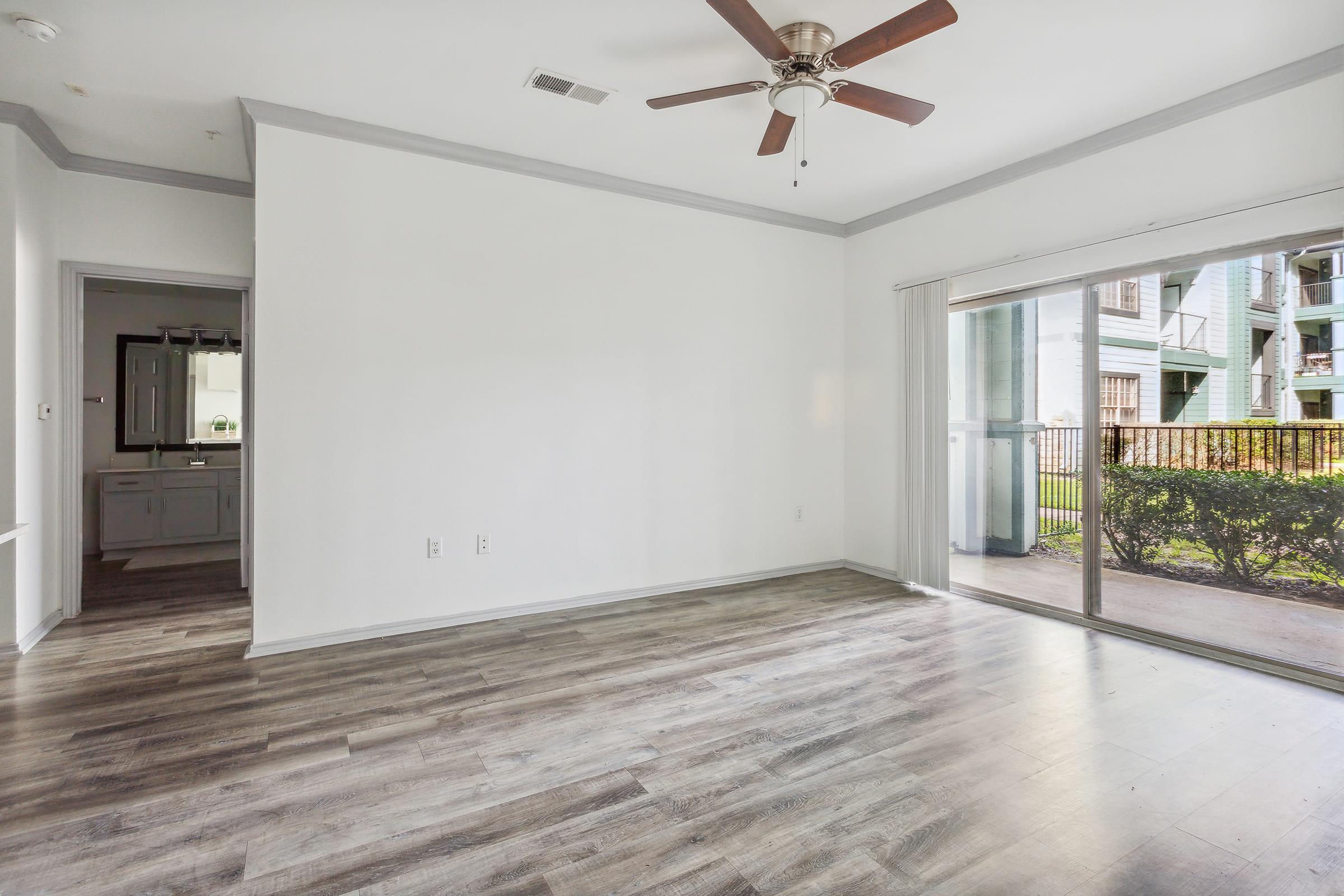 Spacious interior of a living room featuring light-colored walls, a ceiling fan, and large sliding glass doors that lead to an outdoor area. The floor is covered with light wood-like laminate, and there is an open doorway visible leading to another room.