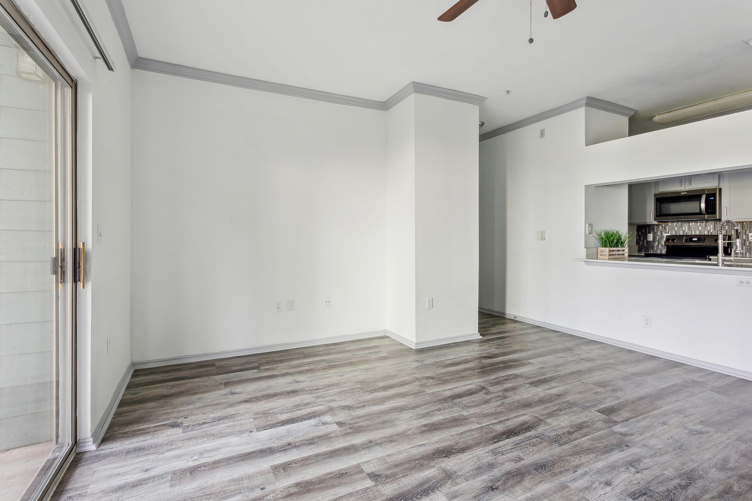 A spacious, empty living area with light-colored walls and a ceiling fan. The floor features wood-like laminate, and there is a small kitchen area visible, including a microwave and modern appliances. A sliding glass door leads to the exterior, allowing natural light to enter the room.
