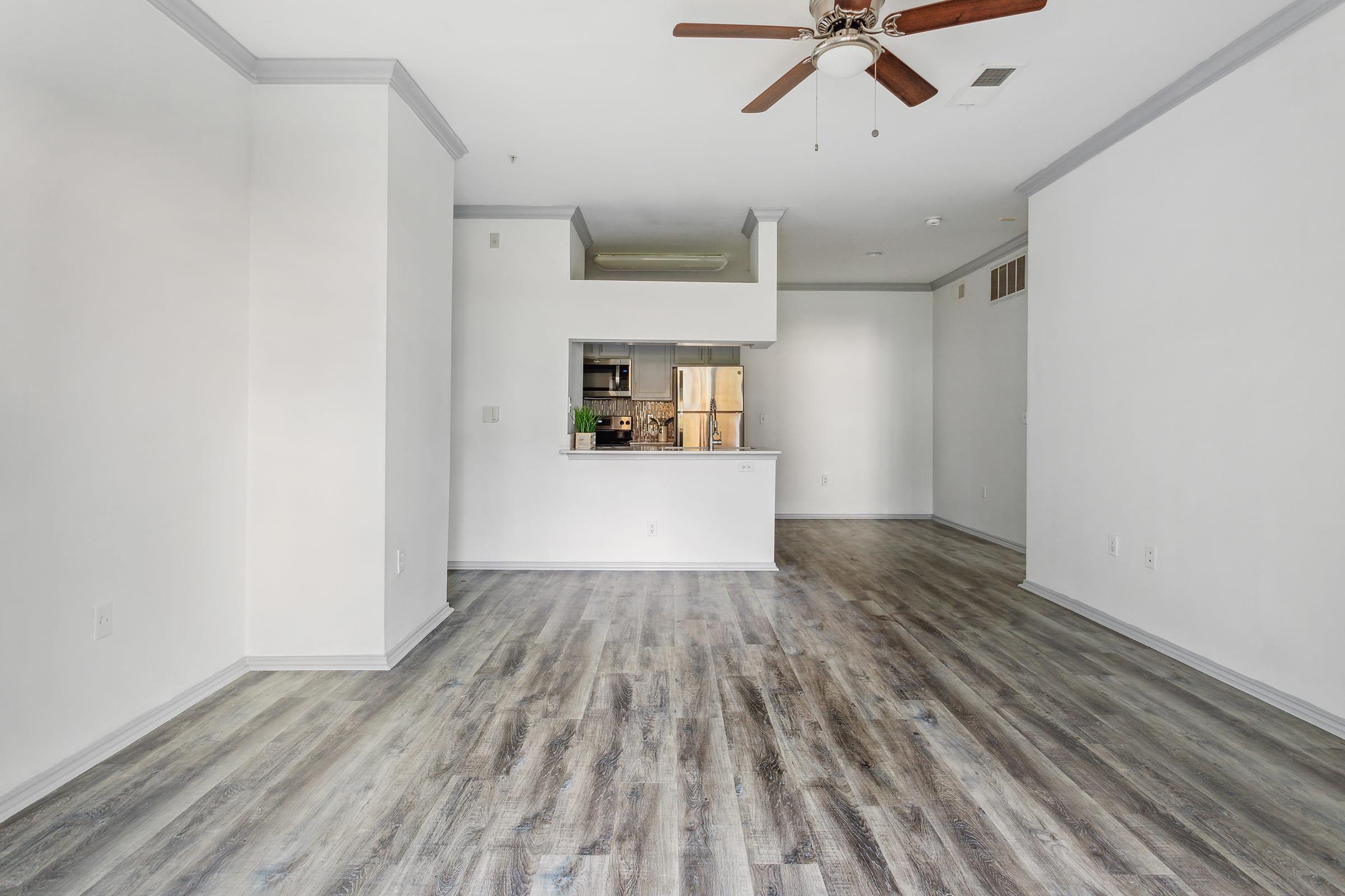 An empty, well-lit living space with light gray walls and wooden flooring. A ceiling fan is visible, and a kitchen area can be seen in the background. The layout is open and spacious, creating a modern atmosphere.