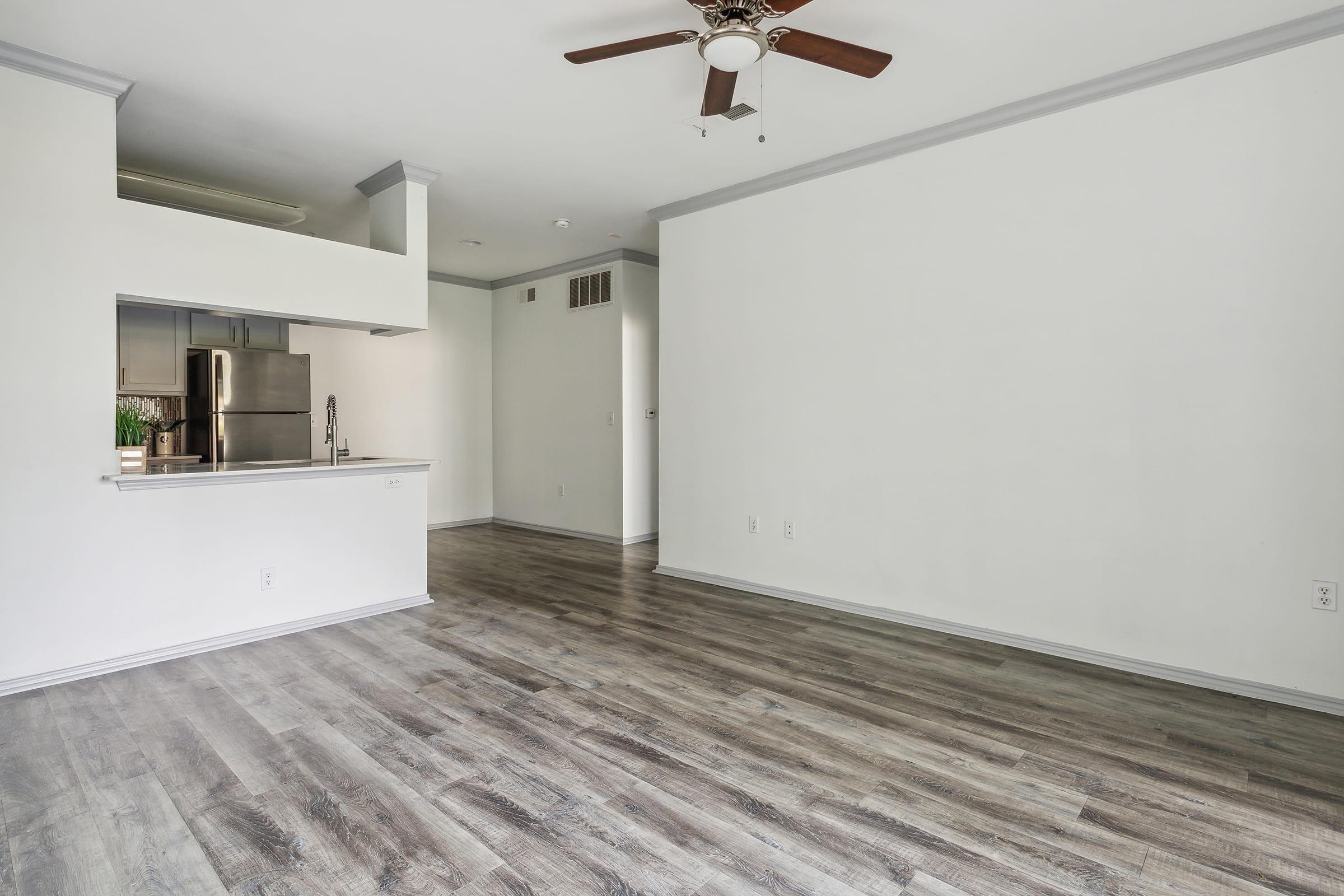 Spacious, empty living area featuring laminate flooring, a ceiling fan, and a minimalist design. A kitchen area is visible with stainless steel appliances and a bar countertop, separated by an open layout. The walls are painted in light colors, creating a bright and airy atmosphere.