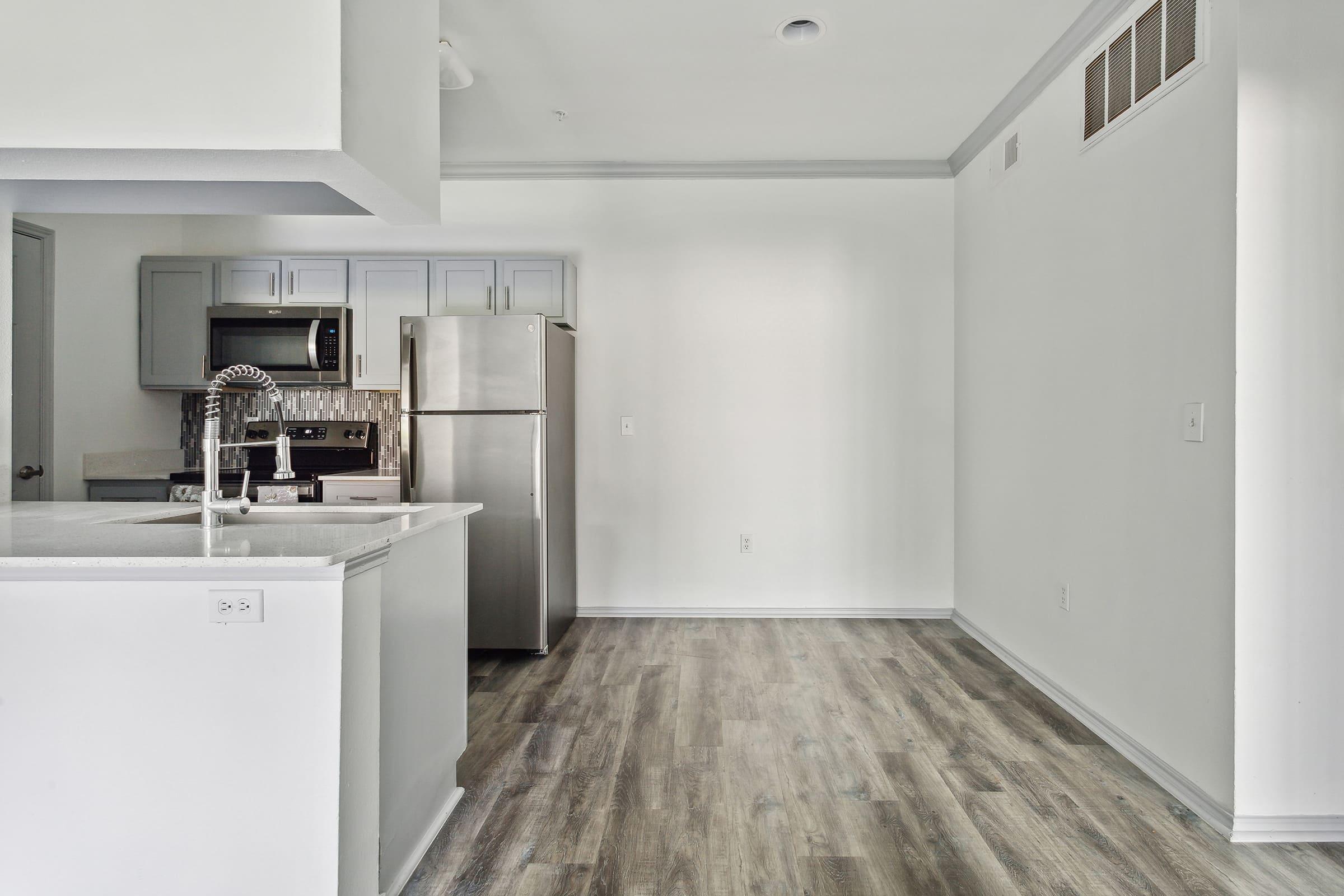 A modern kitchen featuring light gray cabinets, stainless steel refrigerator, and a sleek countertop. The space has an open layout with laminate wood flooring and minimalistic decor. Natural light fills the area, highlighting the clean and stylish design.
