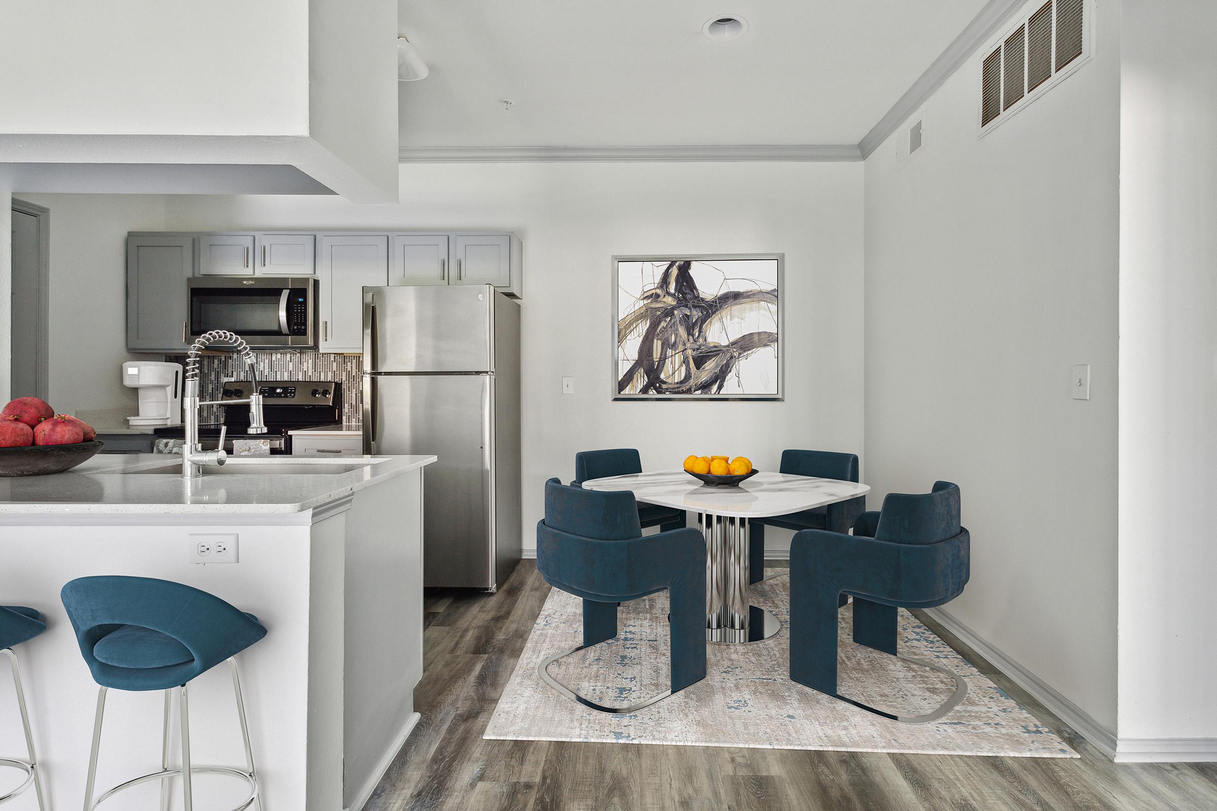 A modern kitchen and dining area featuring stainless steel appliances, a marble dining table surrounded by blue upholstered chairs, and a framed abstract artwork on the wall. A light rug adds warmth to the space, while a bar with stools is positioned near the kitchen.
