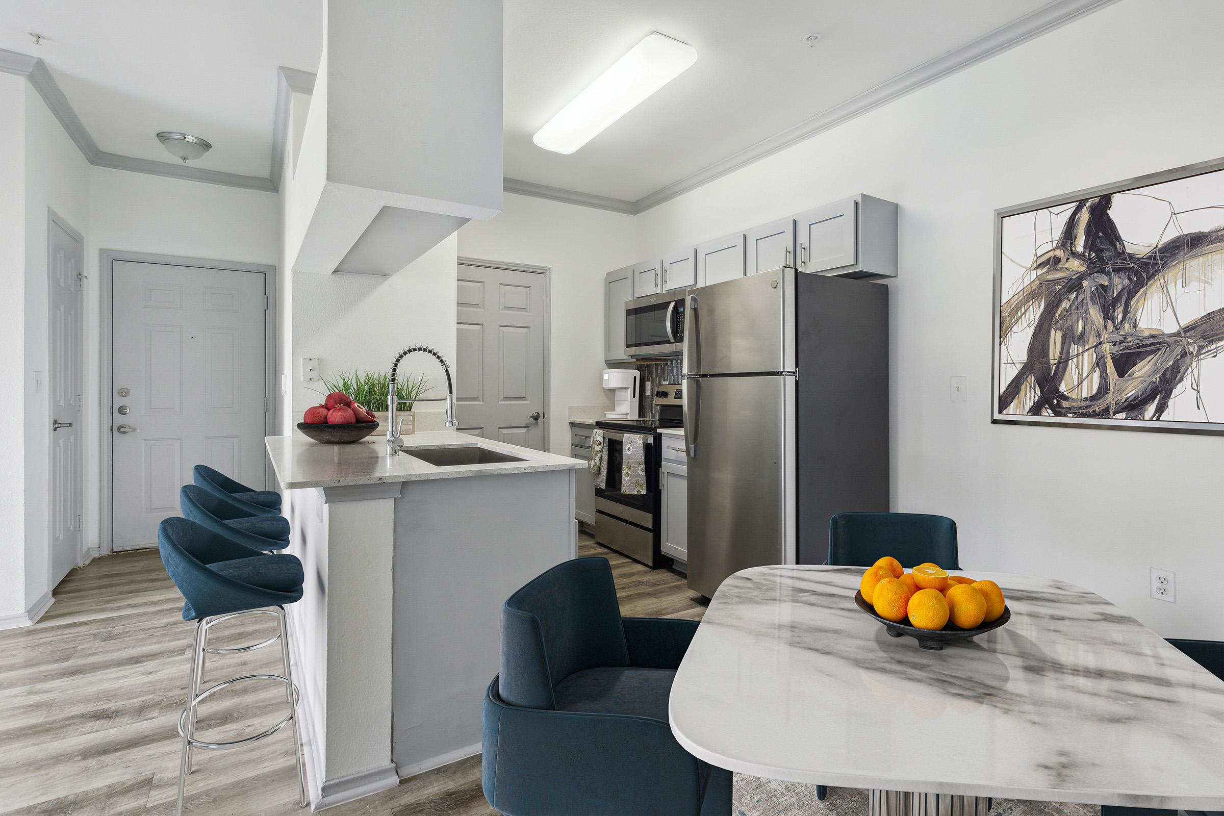 A modern kitchen and dining area featuring a round marble table with a fruit bowl, blue upholstered chairs, stainless steel appliances, and a stylish wall art piece. The space is bright with natural light and has a clean, contemporary design.