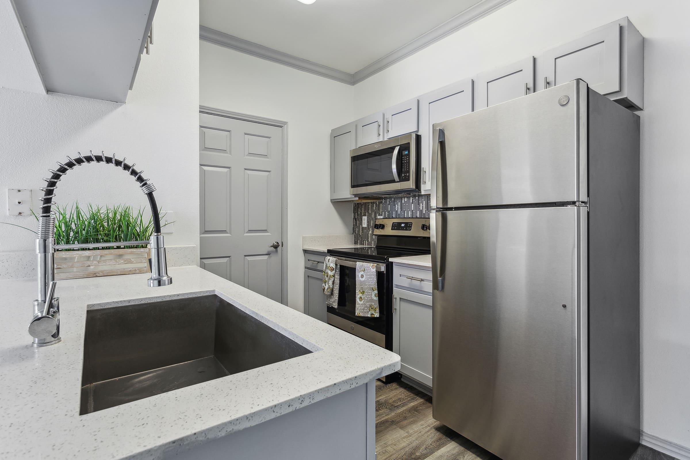 Modern kitchen featuring gray cabinetry, stainless steel appliances including a refrigerator and microwave, a sleek sink with a pull-down faucet, and a decorative backsplash. Natural light illuminates the space, highlighting the stylish countertop and a small potted plant on the counter.