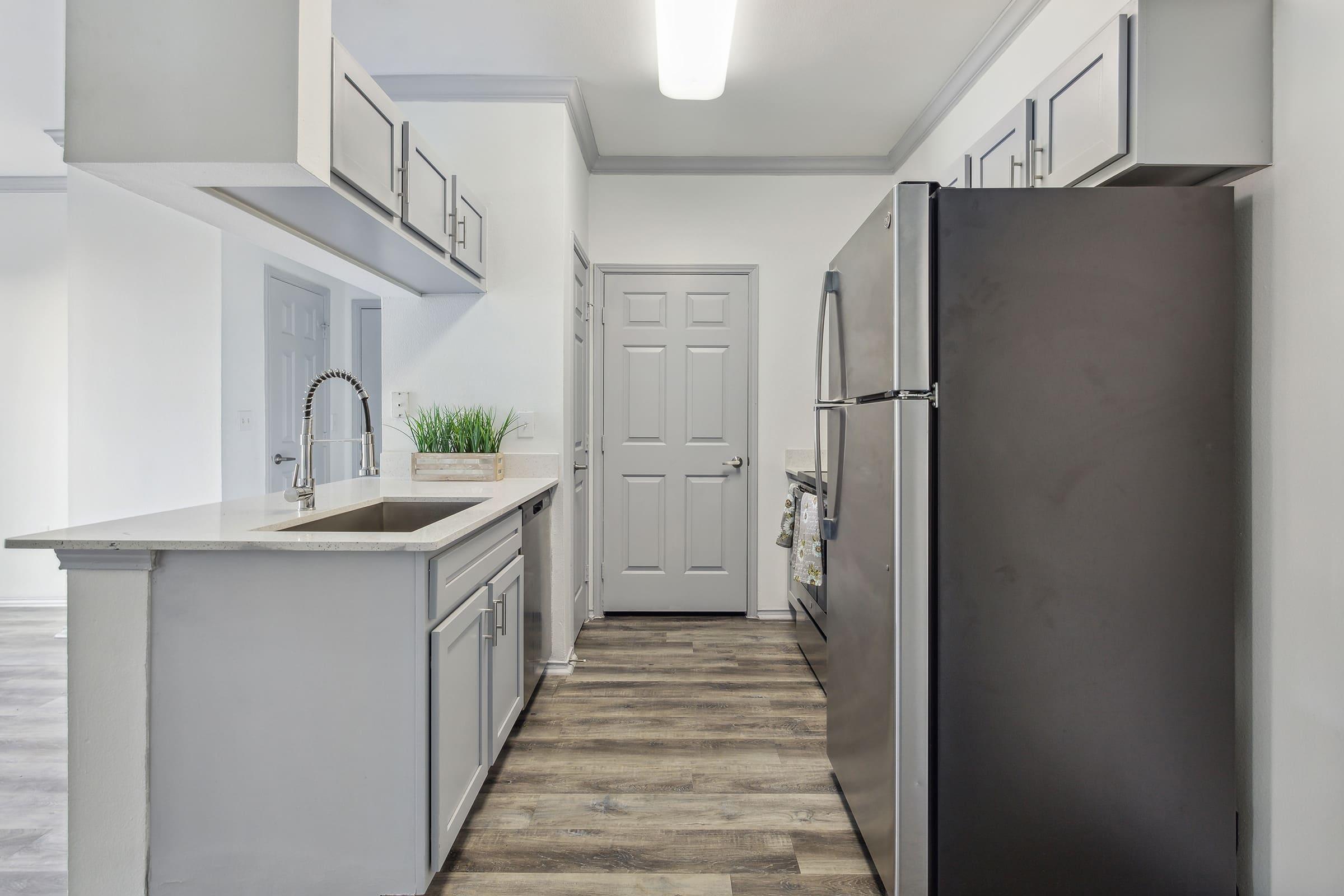 Modern kitchen featuring gray cabinetry, a stainless steel refrigerator, and a countertop with a small plant. The design includes an open layout with neutral-colored walls and wooden flooring, creating a bright and spacious atmosphere. A closed door leads to another area beyond the kitchen.