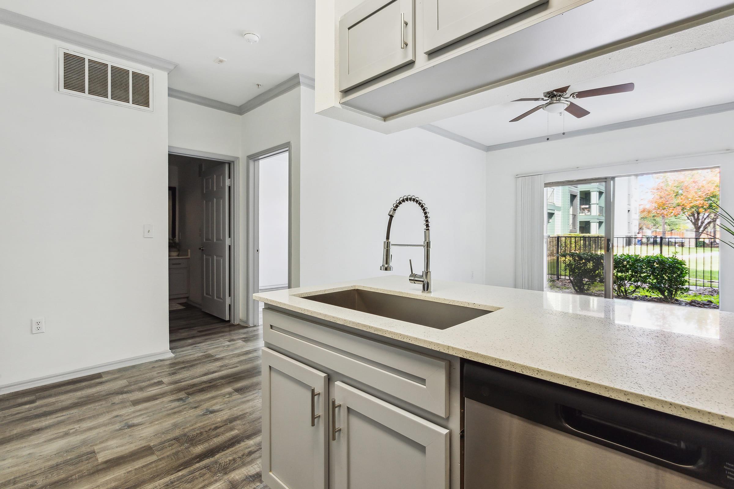 Modern kitchen with a stainless steel sink and faucet, light gray cabinetry, and a dishwasher. In the background, a view of an open living area with large windows overlooking a landscaped yard. The room features wooden flooring and a ceiling fan. A doorway leads to another room. Bright and airy atmosphere.