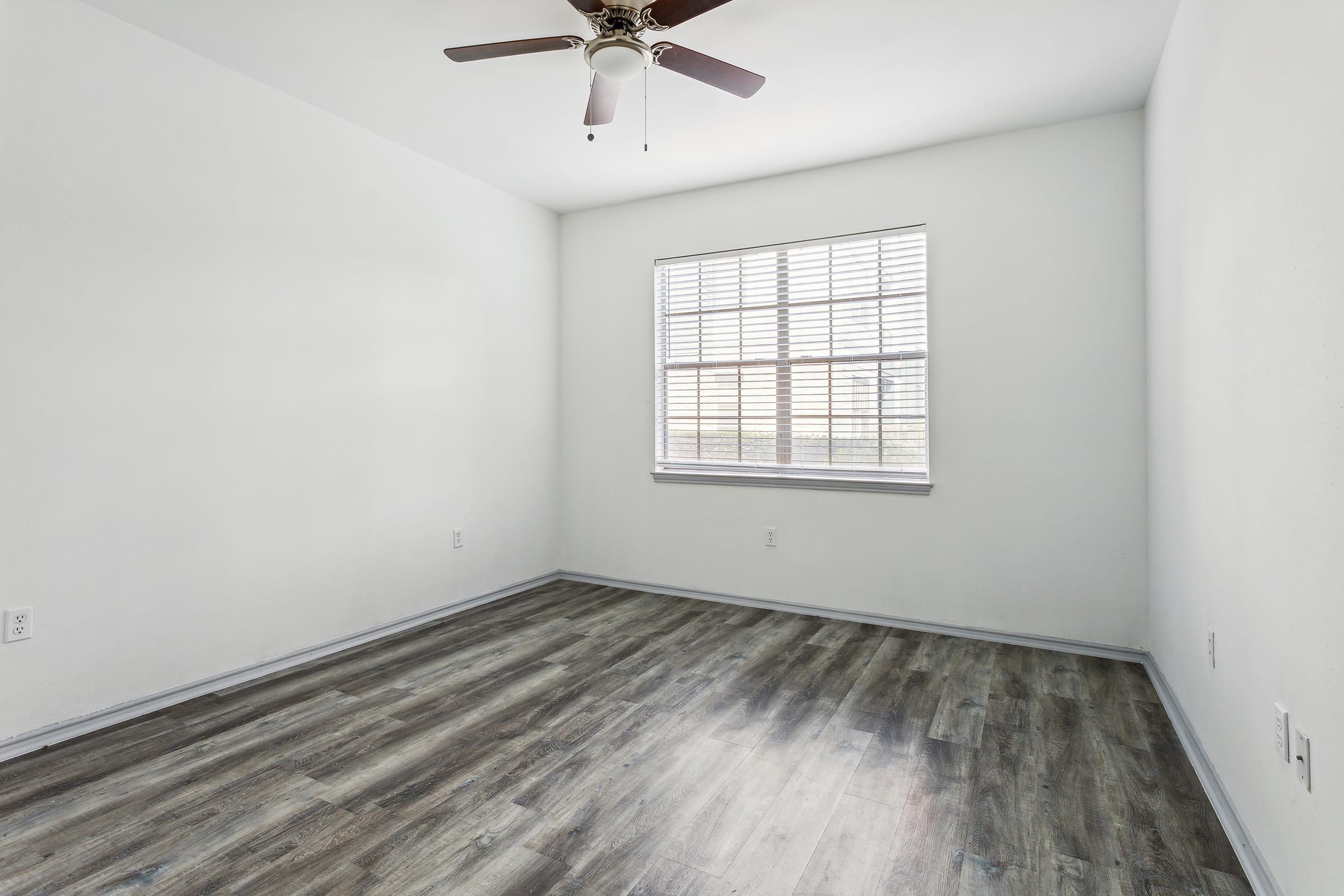 Empty room with light-colored walls and a ceiling fan. There is a window with blinds allowing natural light to enter, and the floor features wood-like laminate. The space appears clean and well-lit, suitable for various uses such as an office or bedroom.