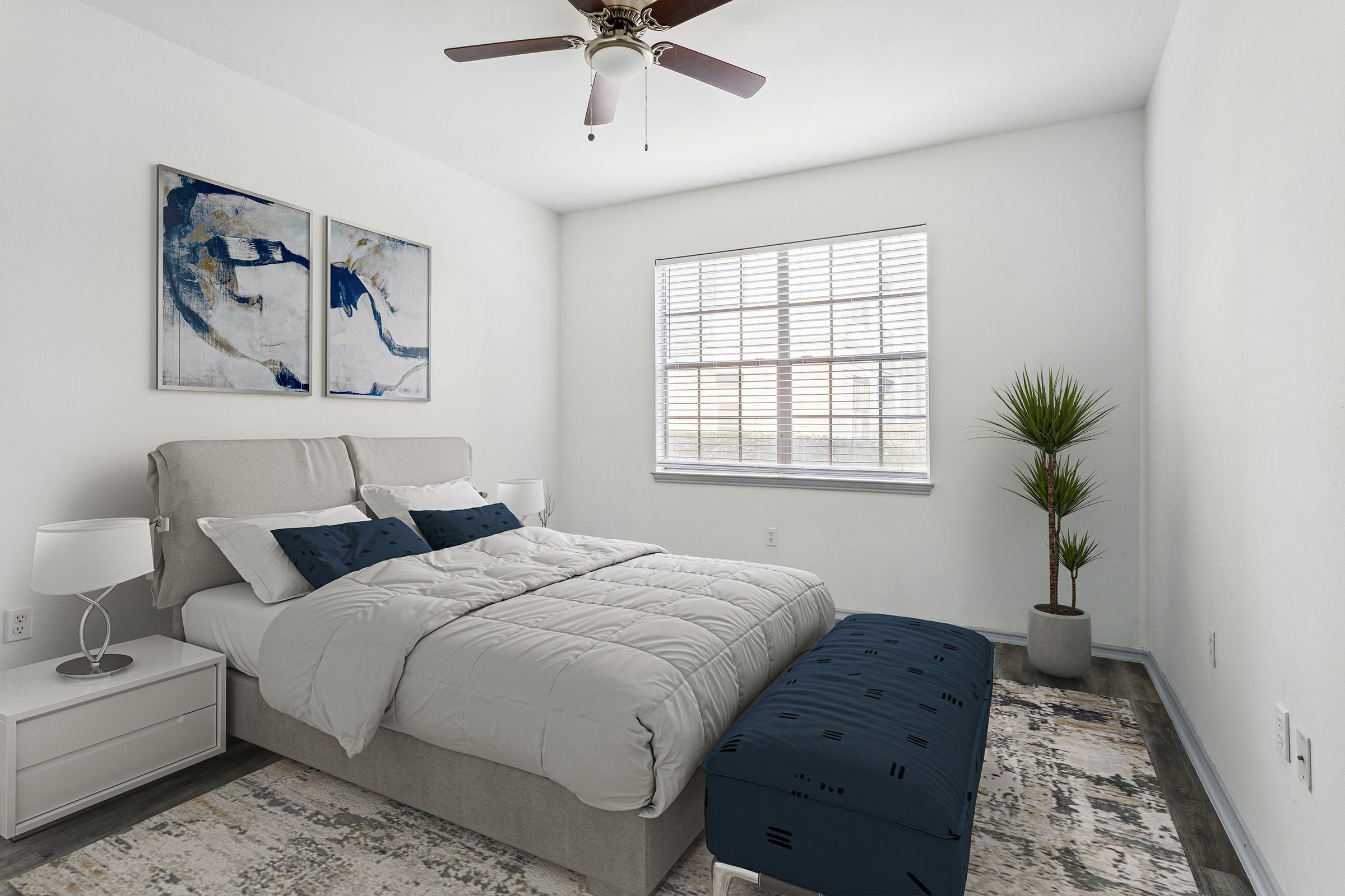 A modern bedroom featuring a light-colored bed with decorative pillows, a cozy bench at the foot, and a nightstand. Two abstract paintings hang on the wall, and a potted plant is situated near the window, which has blinds that let in natural light. The walls are painted white, creating a bright and airy atmosphere.